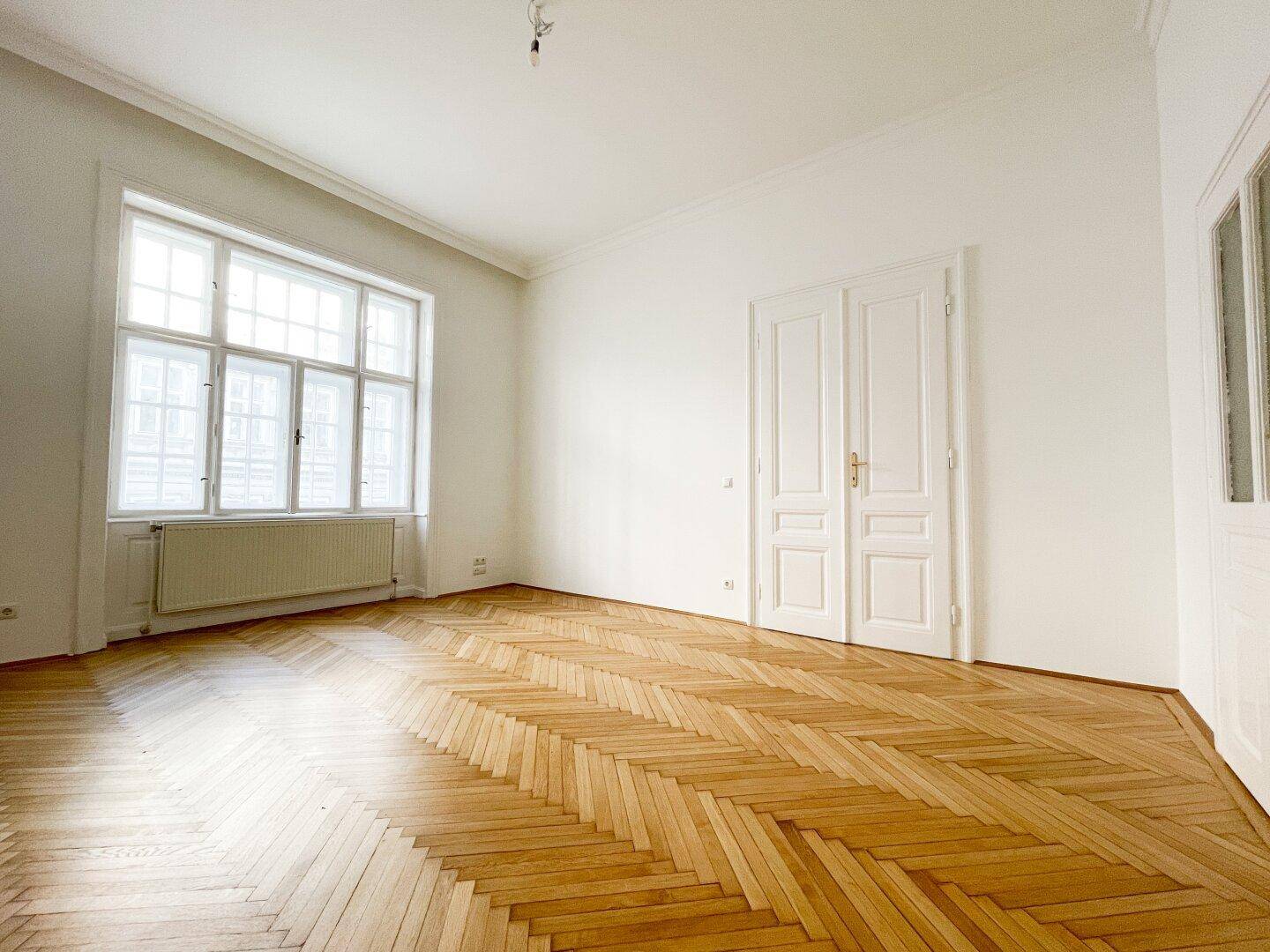 Geräumiges, helles Zimmer mit Fischgrätparkett und großen Fenstern, die viel Licht spenden.