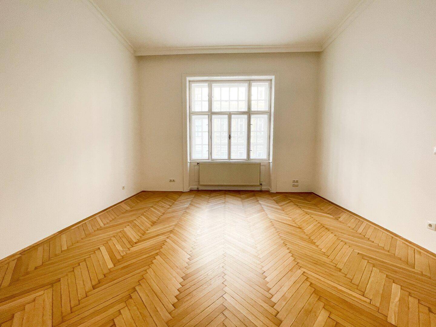 Leeres Zimmer mit Fischgrätparkett und einem großen Fenster, das den Raum erhellt.