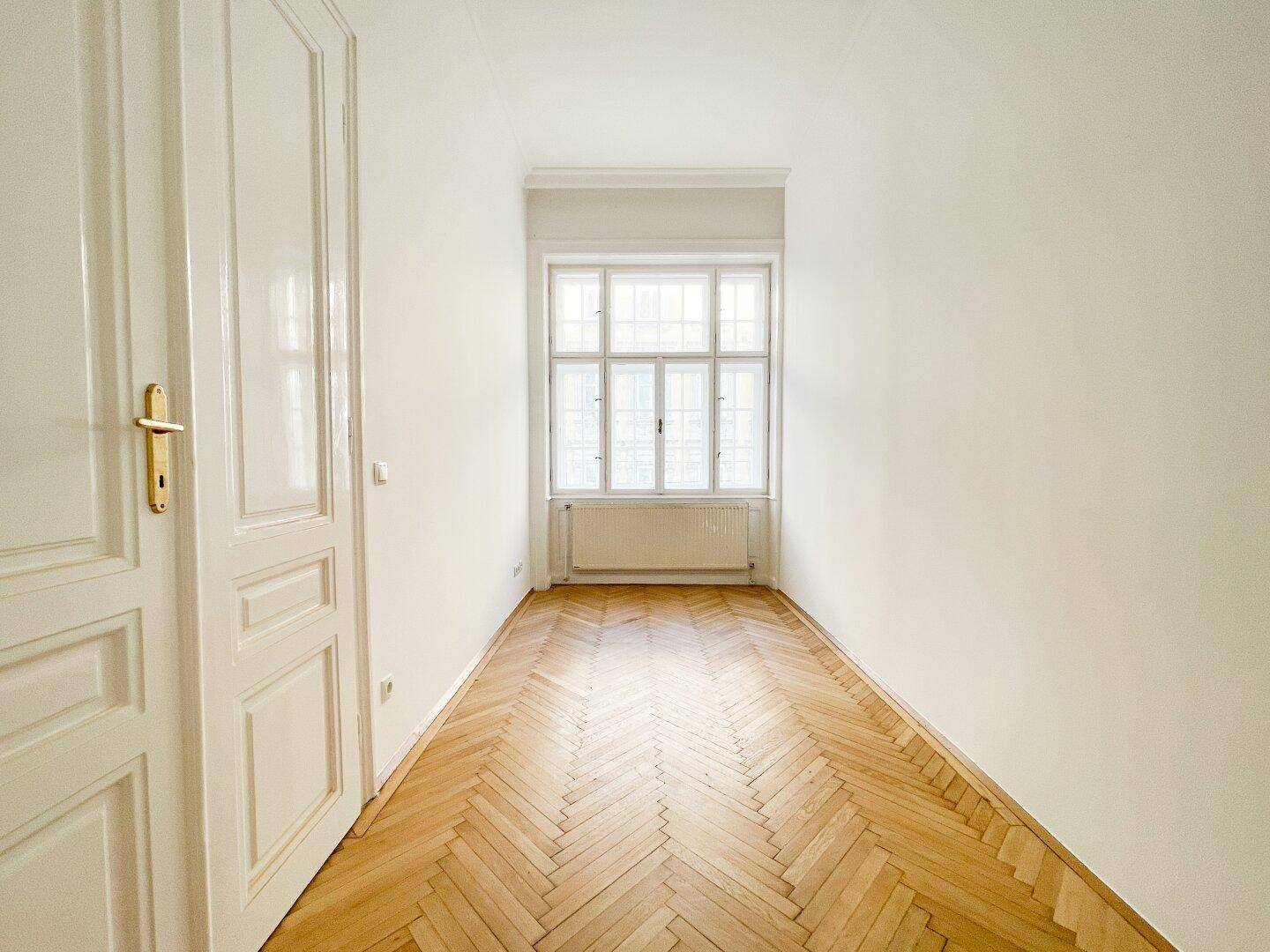 Schmaler, heller Raum mit Fischgrätparkett und einem Fenster am Ende, ideal für ein Büro.