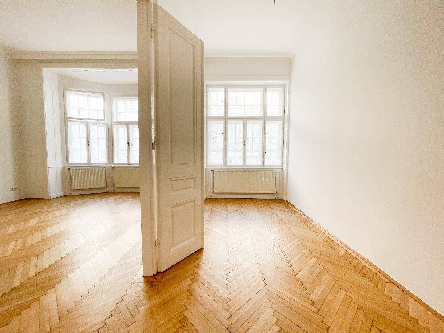 Helles Zimmer mit Fischgrätparkett und großen Fenstern, die den Raum mit Licht füllen.