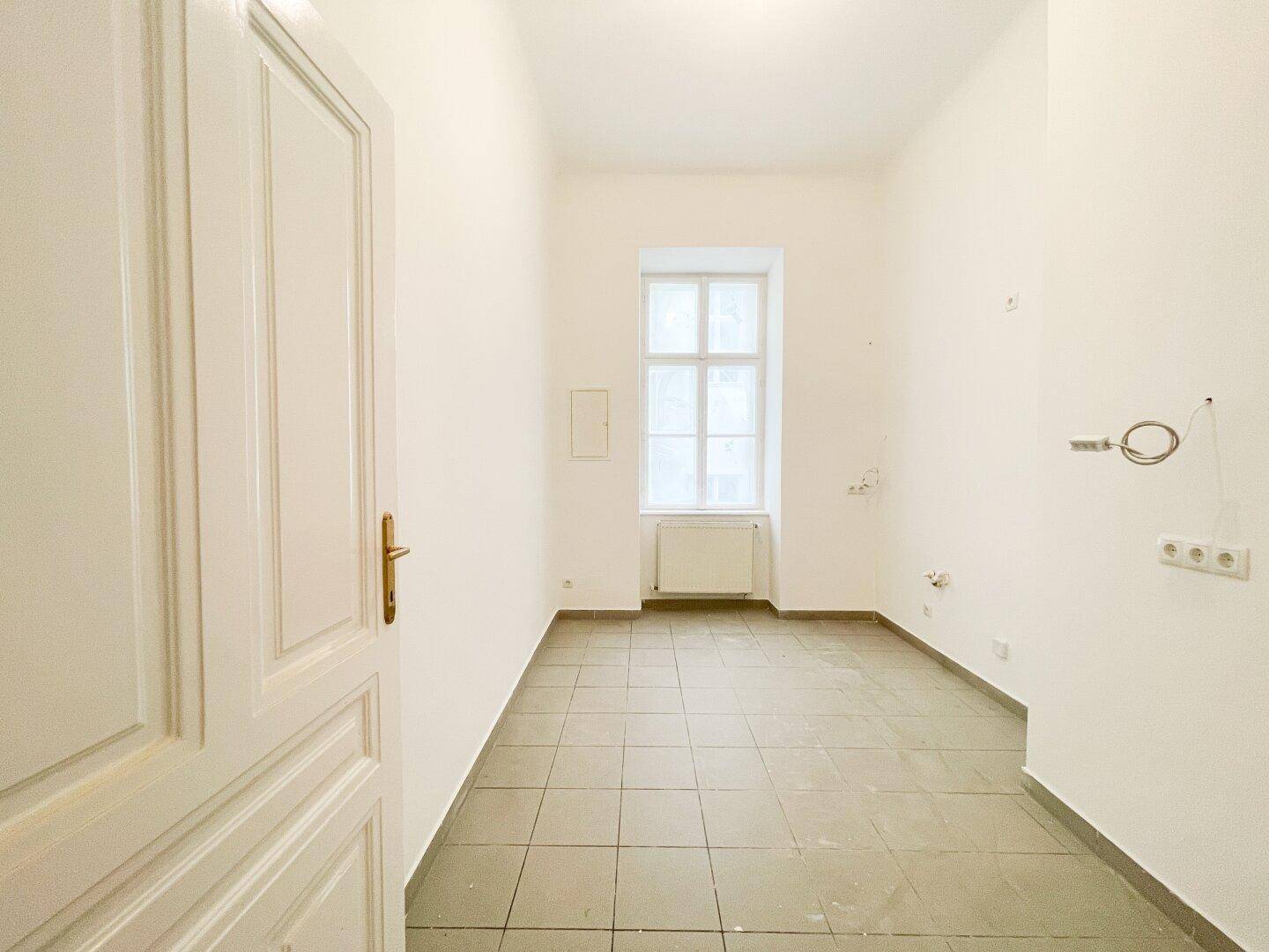 Leerer Raum mit Fliesenboden und vorbereiteten Anschlüssen für eine zukünftige Küche.