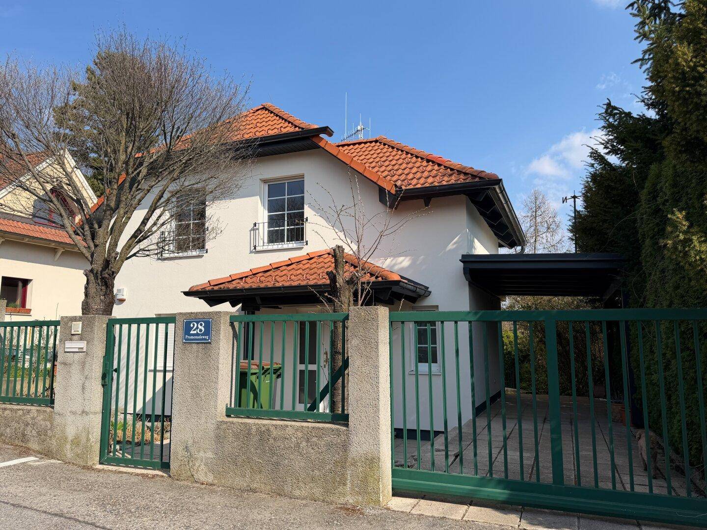 Vorderansicht eines Einfamilienhauses mit grünem Zaun, Carport und gepflegter Umgebung.
