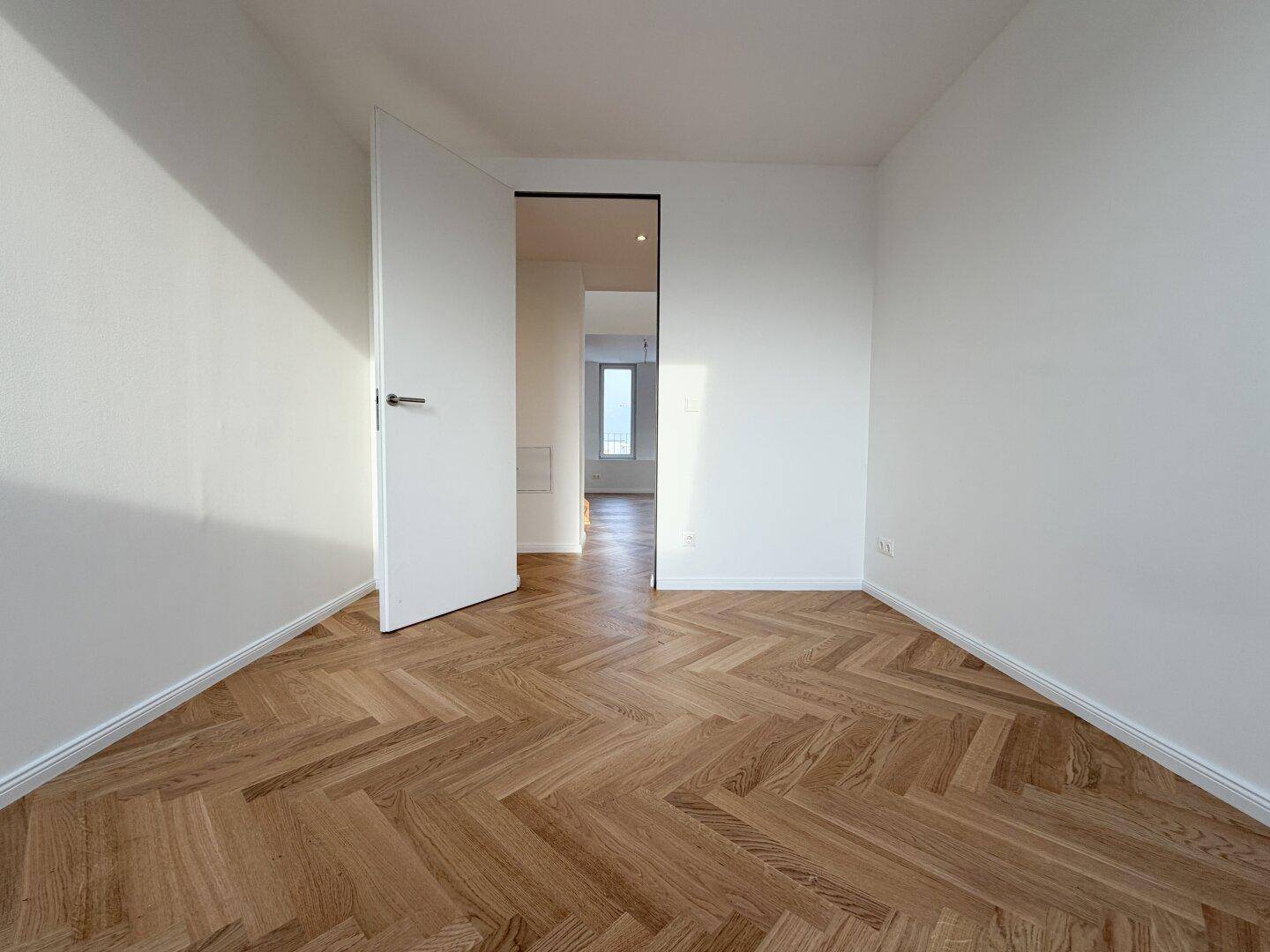 Heller Raum mit Fischgrätparkett und offener Tür, die den Blick in den Flur freigibt.