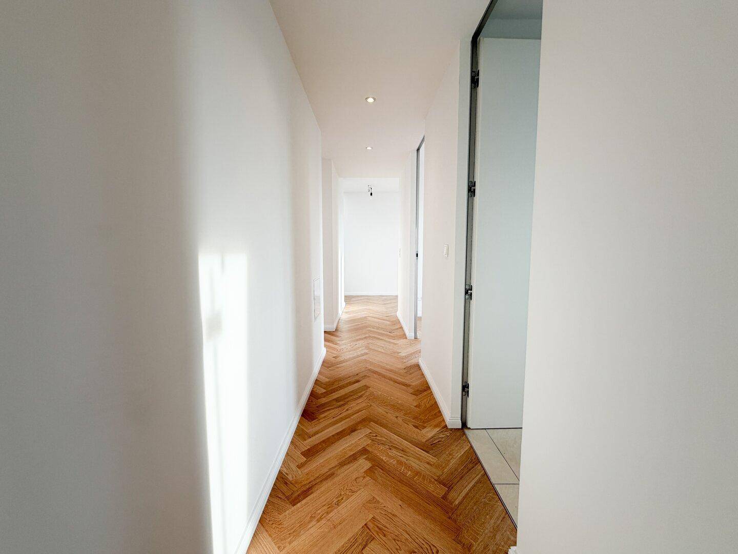 Langer, heller Flur mit Fischgrätparkett und Einbaustrahlern in der Decke.