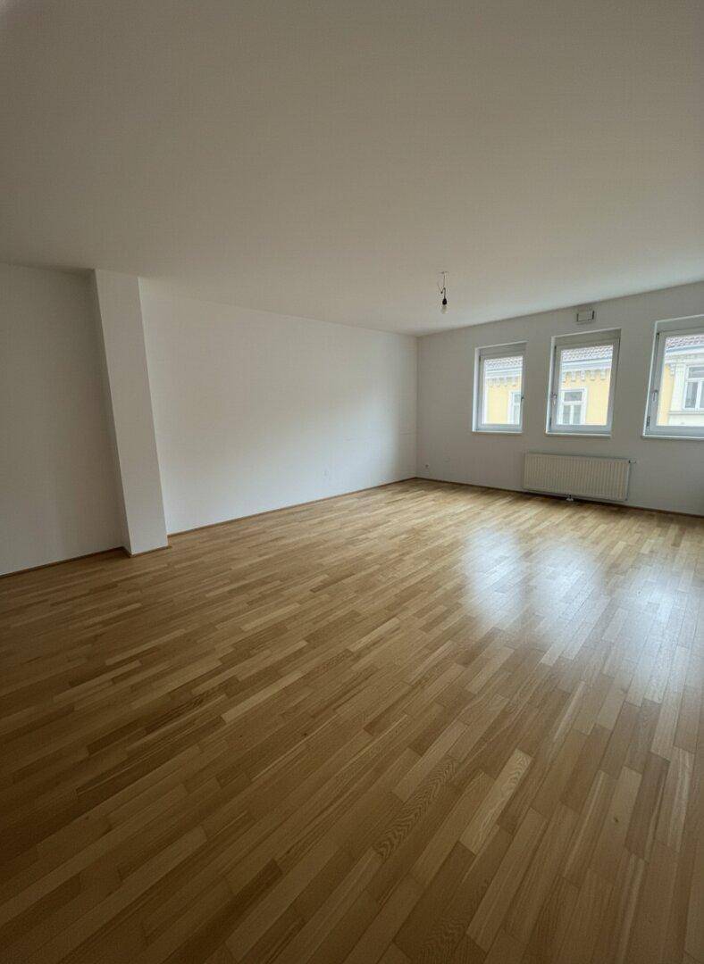 Geräumiges, leeres Wohnzimmer mit hellem Parkettboden und drei großen Fenstern.