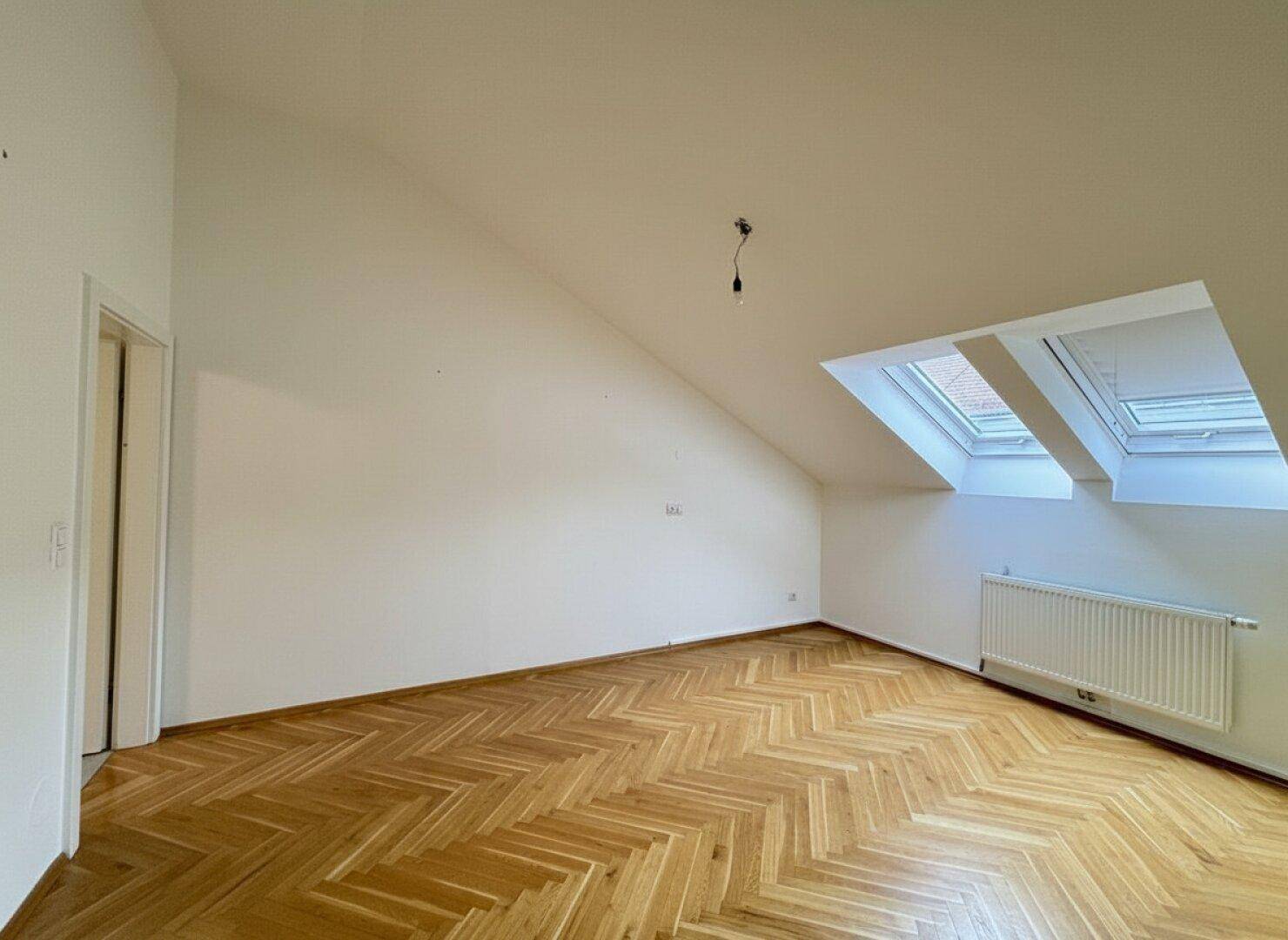 Geräumiges Dachgeschosszimmer mit Parkettboden, schräger Decke und zwei Dachfenstern.