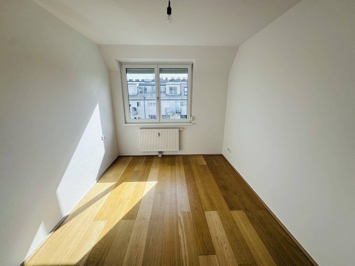 Helles Zimmer mit Holzparkett, einem Fenster und viel Tageslicht.