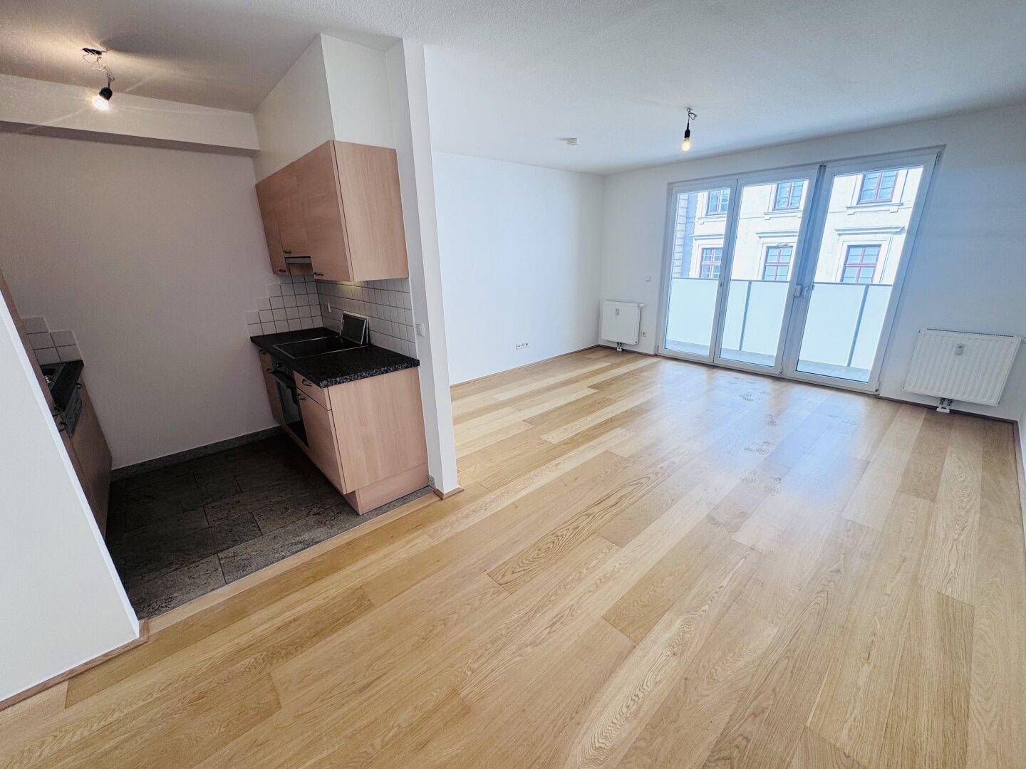 Offener Wohnbereich mit Kochnische, Holzparkett und Zugang zum Balkon.