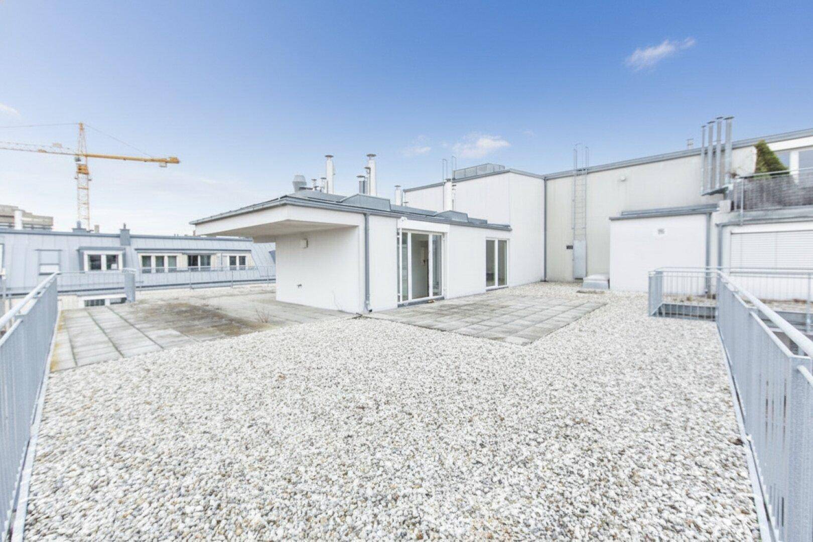 Weitläufige Dachterrasse mit Kiesfläche und Fliesenbereich, umgeben von modernen Gebäudestrukturen und offenem Himmel.