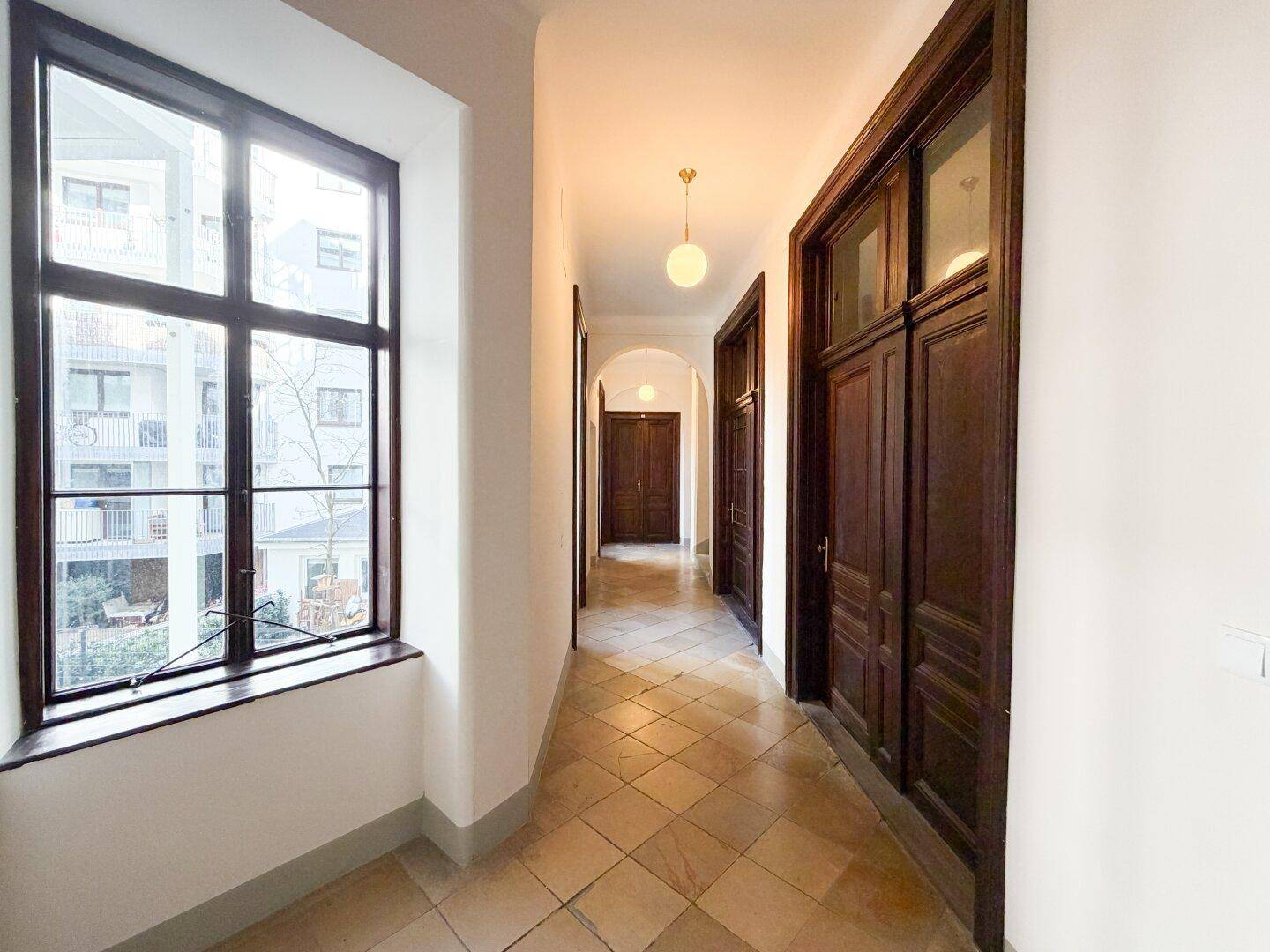 Langer Flur mit Fliesenboden, großen Fenstern und dunklen Holztüren.