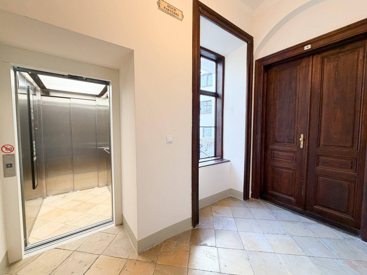 Heller Flur mit Aufzug, Fliesenboden und Holztüren zu den Wohnungen.