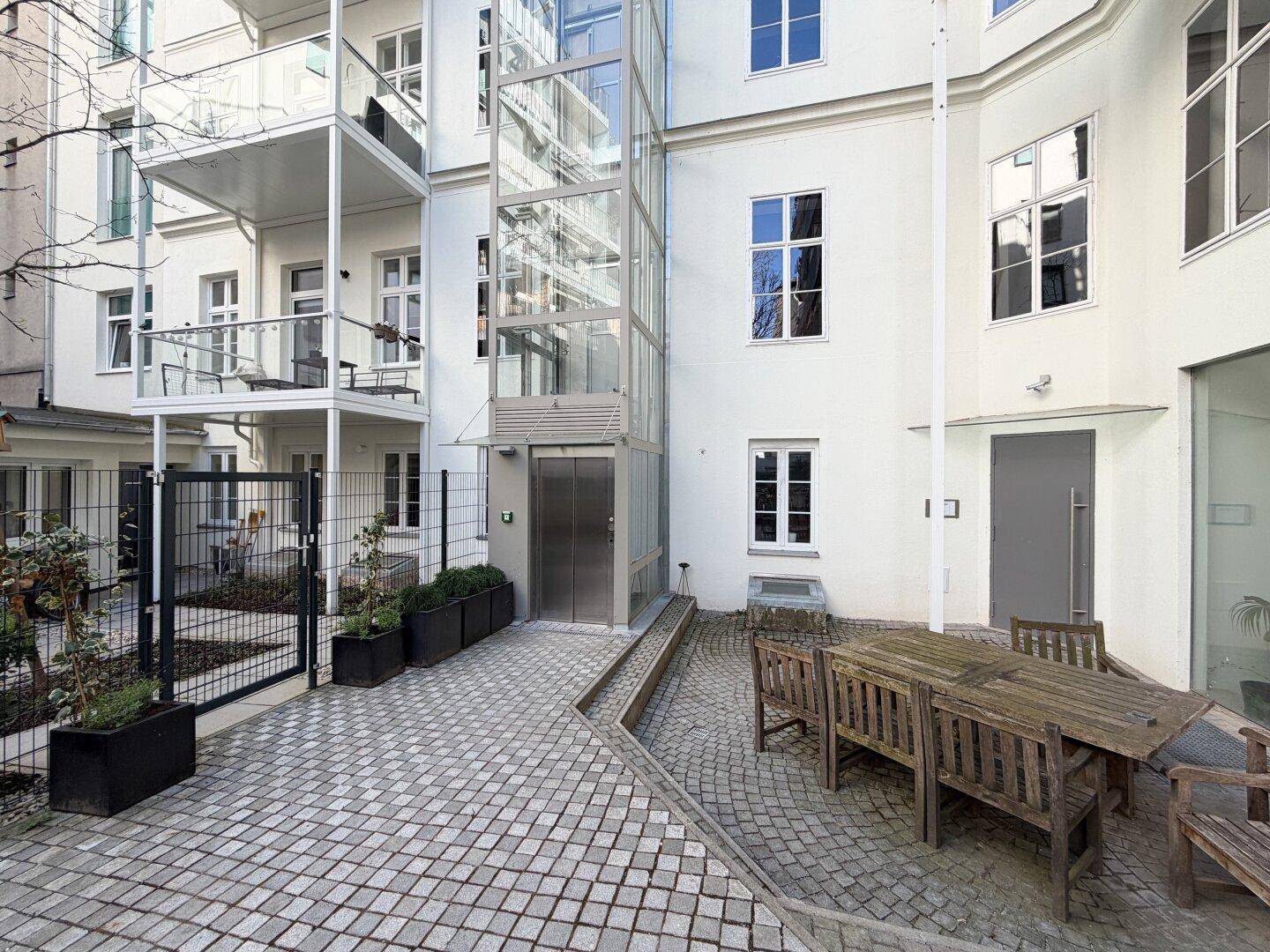 Gepflasterter Innenhof mit Sitzgelegenheiten und Zugang zum modernen Glasaufzug.