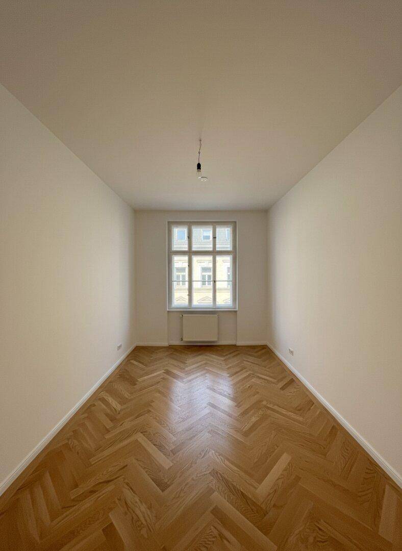 Helles, leeres Zimmer mit Fischgrätparkett und einem großen Fenster.