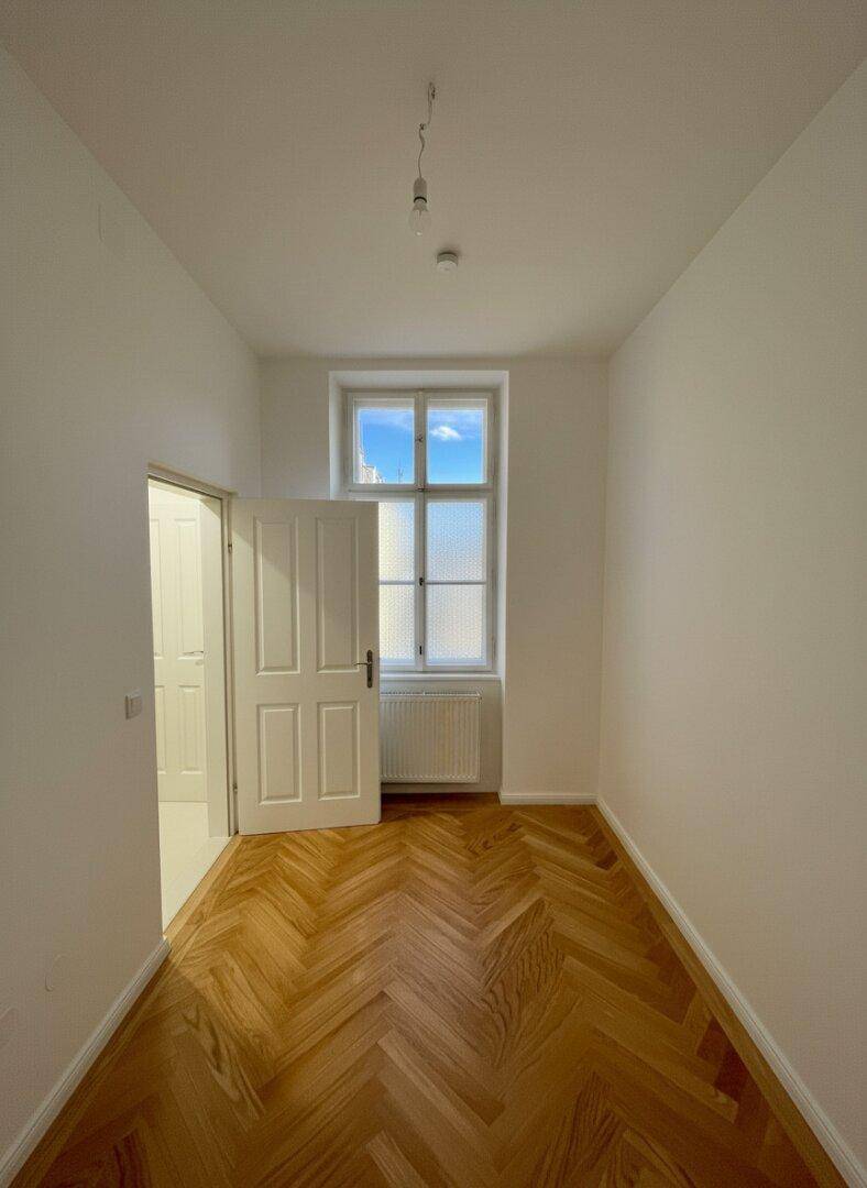 Langes, helles Zimmer mit Holzboden und einem Fenster, das viel Licht hereinlässt.