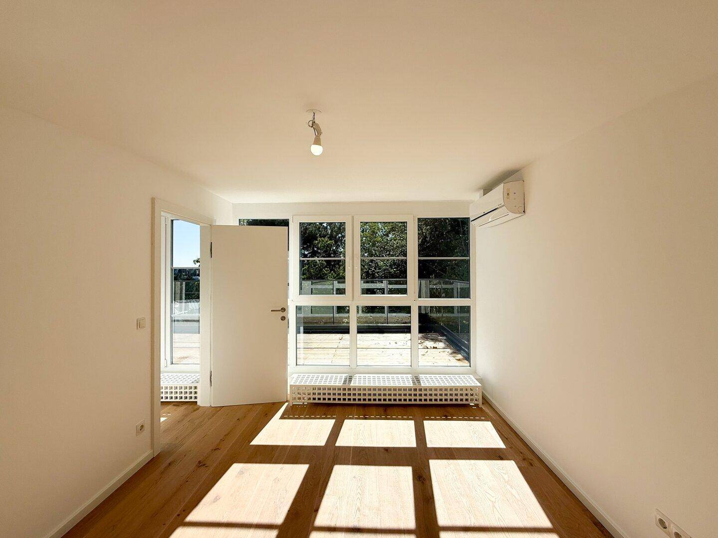 Heller Raum mit Holzboden, großen Fenstern und Zugang zu einem Außenbereich.
