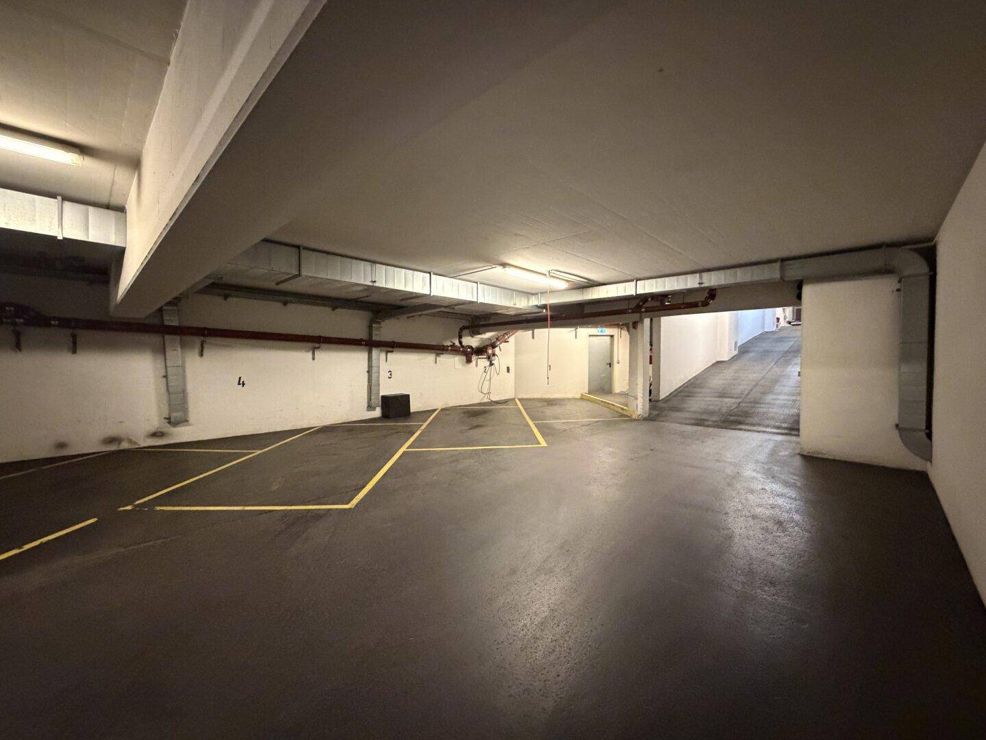 Weitläufige Tiefgarage mit zahlreichen markierten Parkplätzen und sichtbaren Installationen an der Decke und Wänden.