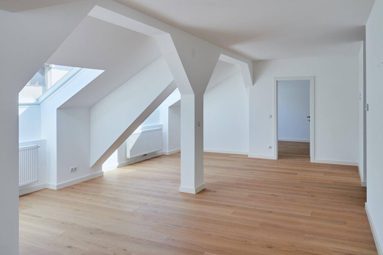 Lichtdurchfluteter Dachgeschossraum mit Holzboden, schrägen Wänden und Zugang zu weiteren Zimmern.