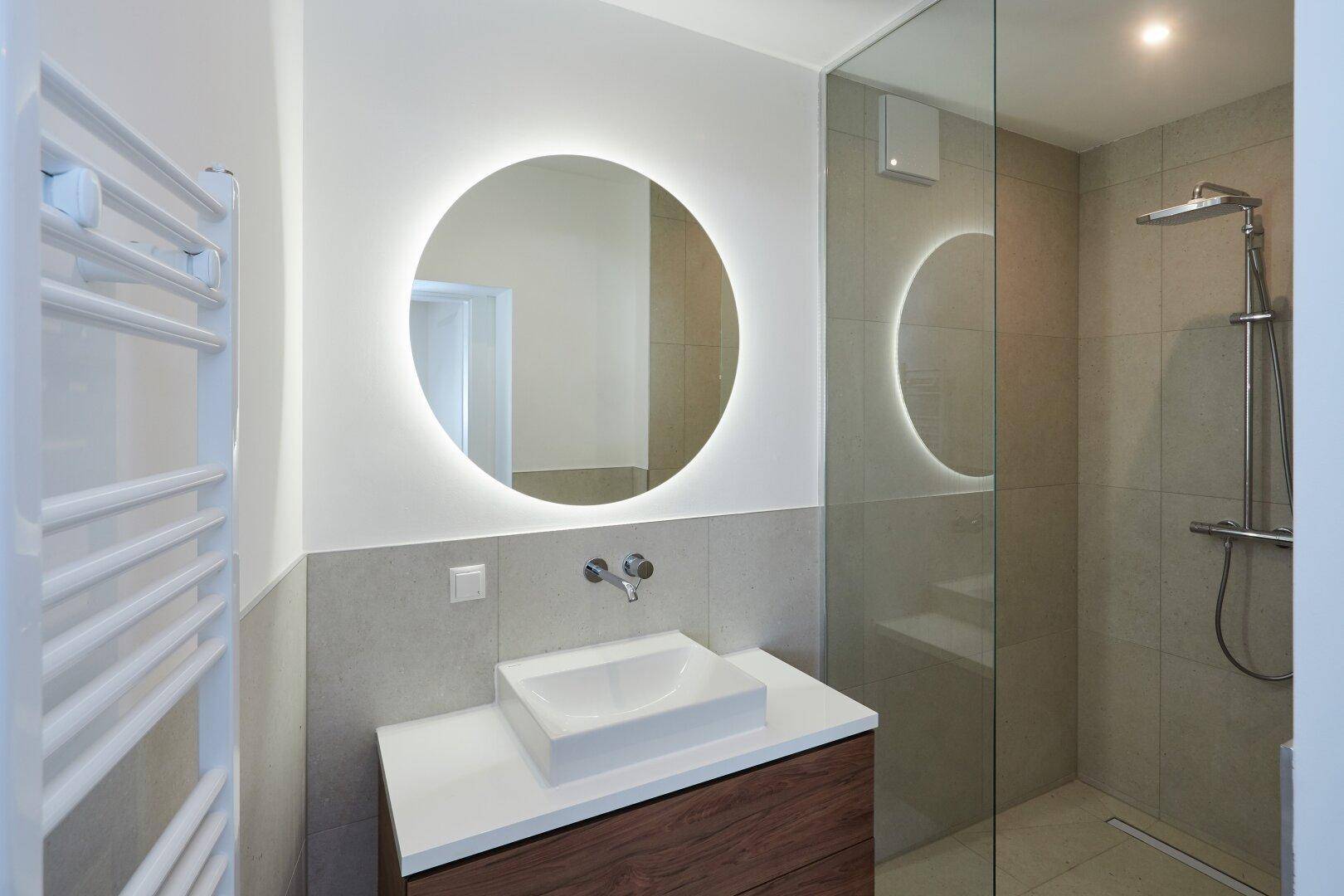 Modernes Badezimmer mit beleuchtetem Spiegel, Einzelwaschbecken und ebenerdiger Dusche.
