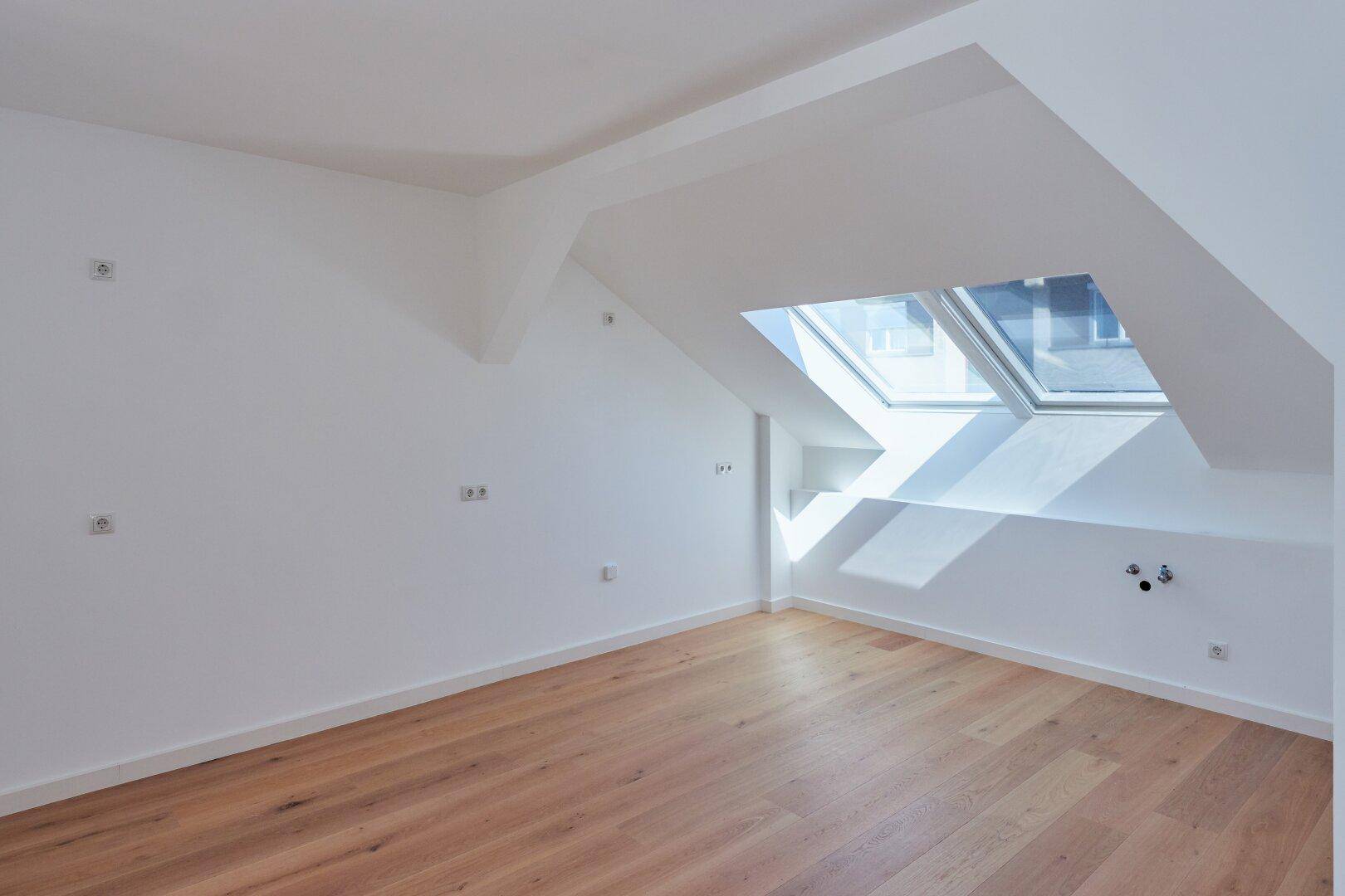 Heller Dachgeschossraum mit Holzboden und schrägen Wänden, beleuchtet durch ein großes Dachfenster.