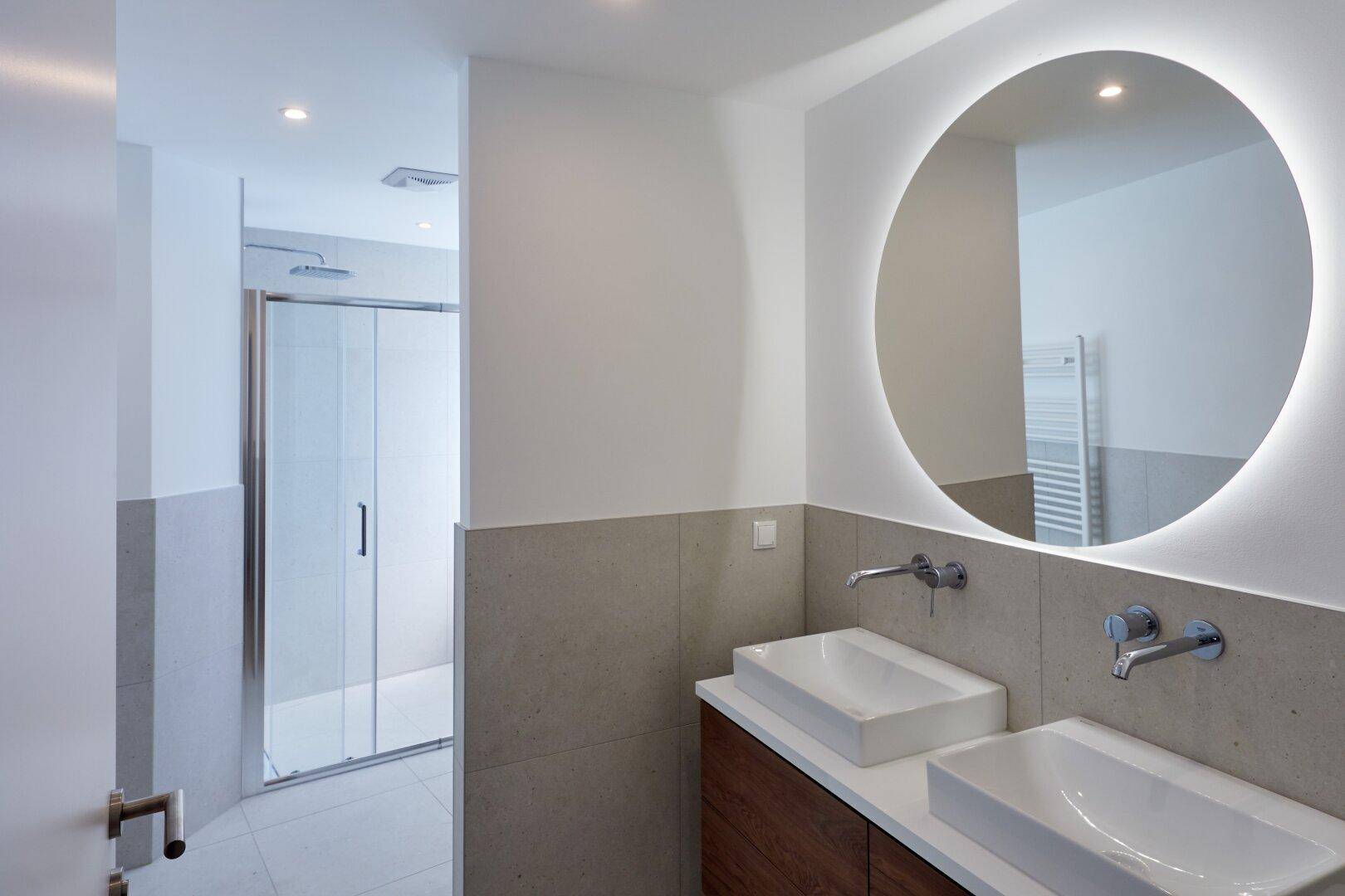 Modernes Badezimmer mit Doppelwaschbecken, beleuchtetem Spiegel und Duschbereich.