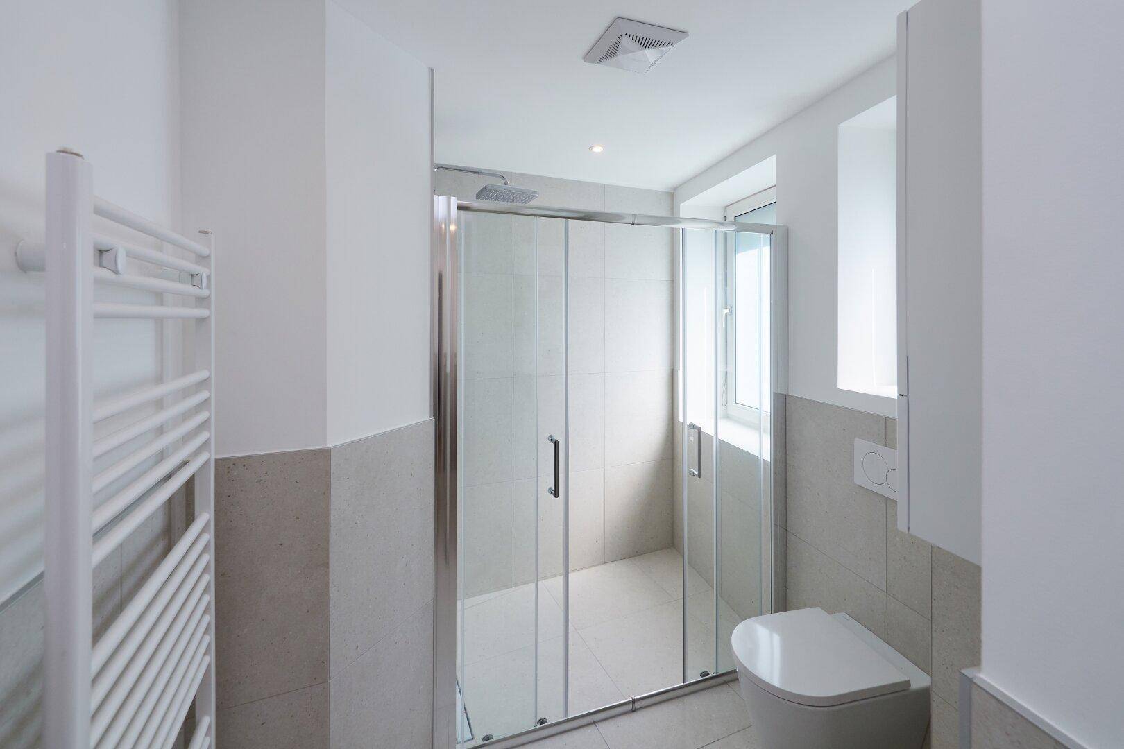 Helles Badezimmer mit Duschkabine, wandmontierter Toilette und Fenster für Tageslicht.