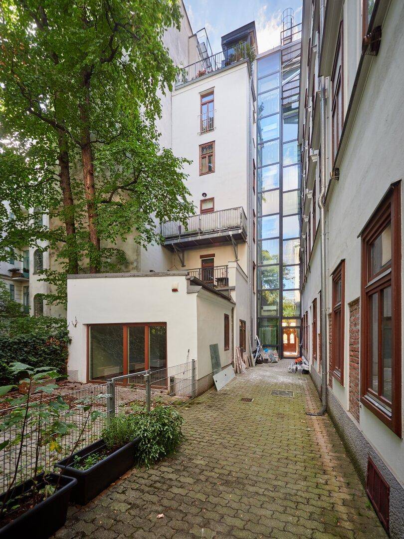 Hinterhofansicht des Gebäudes mit modernem Glasaufzug und angrenzendem Gartenbereich.