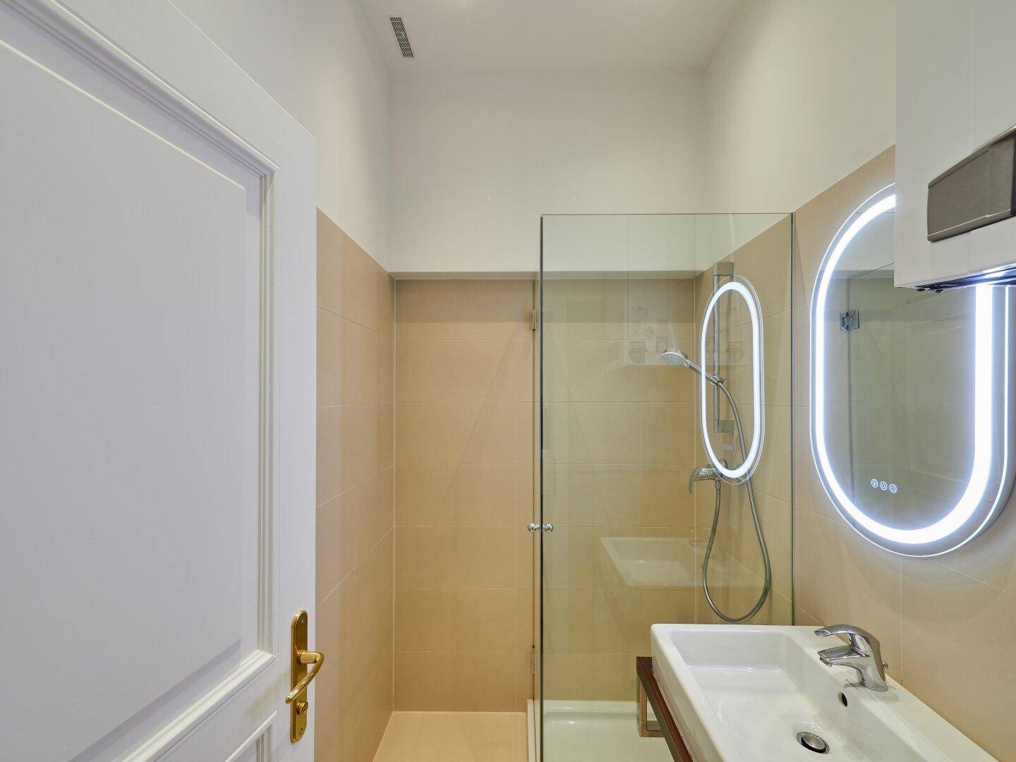 Modernes Badezimmer mit begehbarer Dusche, beleuchtetem Spiegel und beigefarbenen Fliesen.