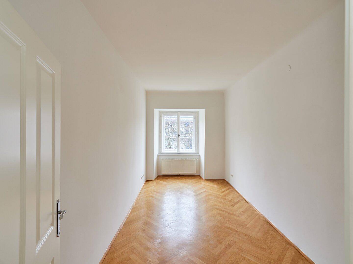 Langes, helles Zimmer mit Fischgrätparkett und einem Fenster, das den Raum mit Licht erfüllt.