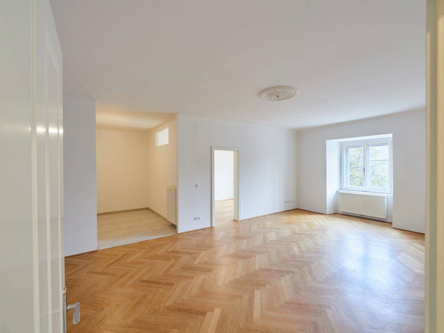 Geräumiges Zimmer mit Fischgrätparkett und Zugang zu einem weiteren Bereich, hell und einladend.