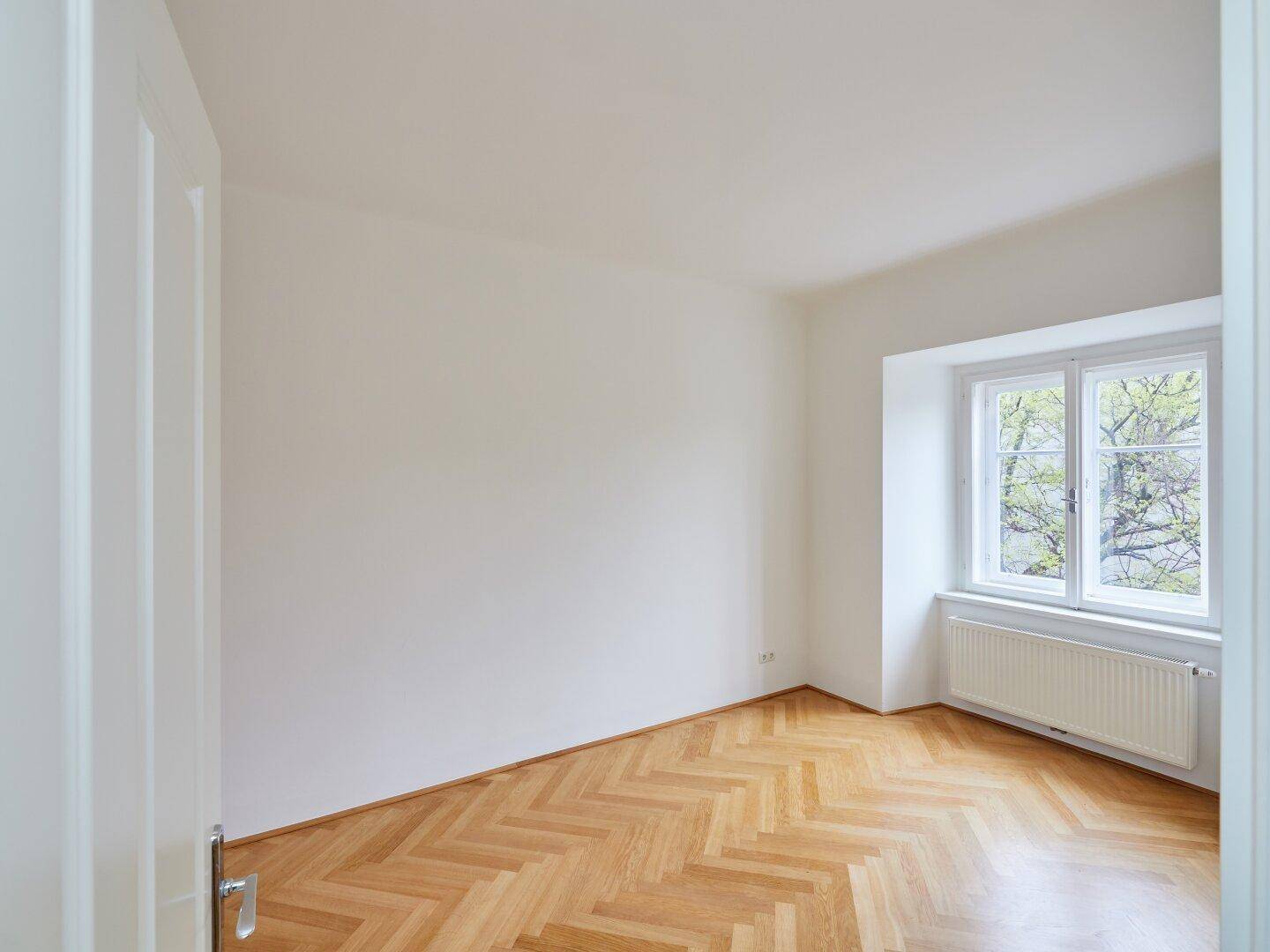 Heller Raum mit Fischgrätparkett und einem Fenster, das einen Blick ins Grüne bietet.