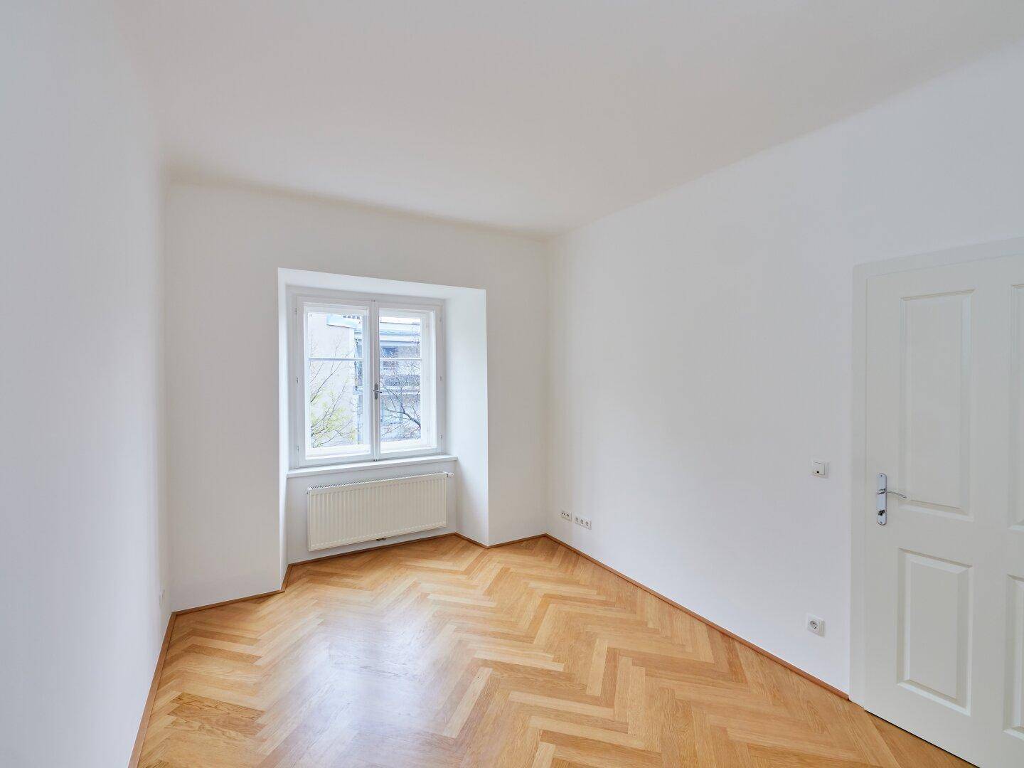 Helles Zimmer mit Fischgrätparkett und einer Tür, die zu einem weiteren Raum führt.