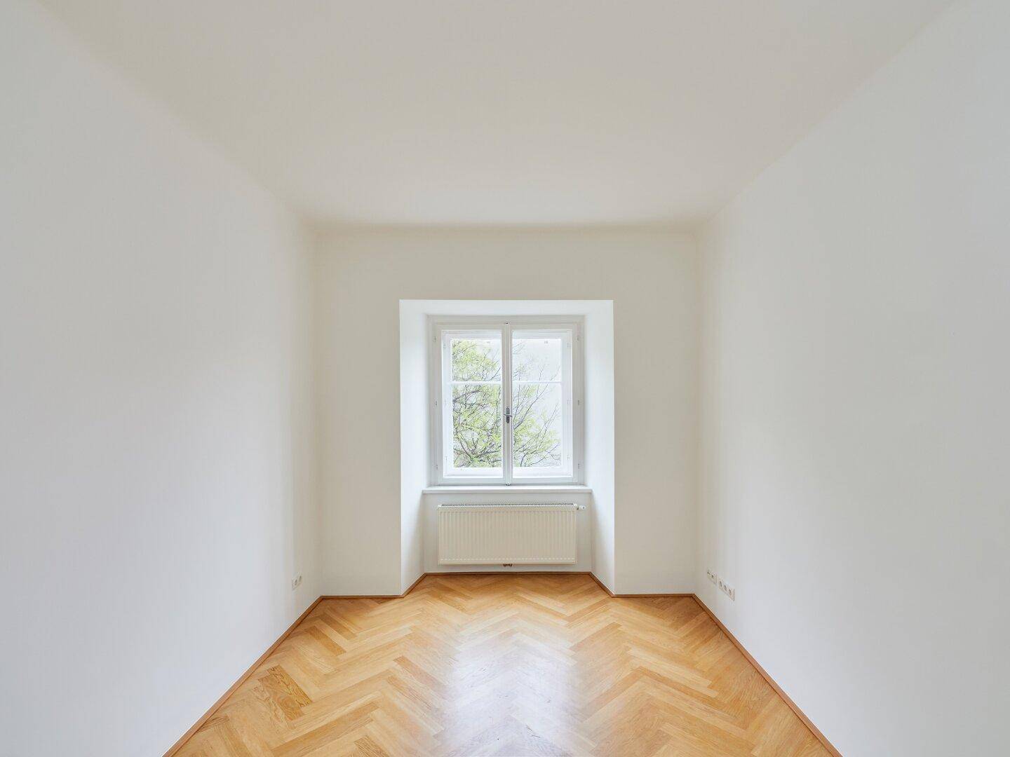Symmetrischer Raum mit Fischgrätparkett und einem zentralen Fenster, das viel Licht spendet.