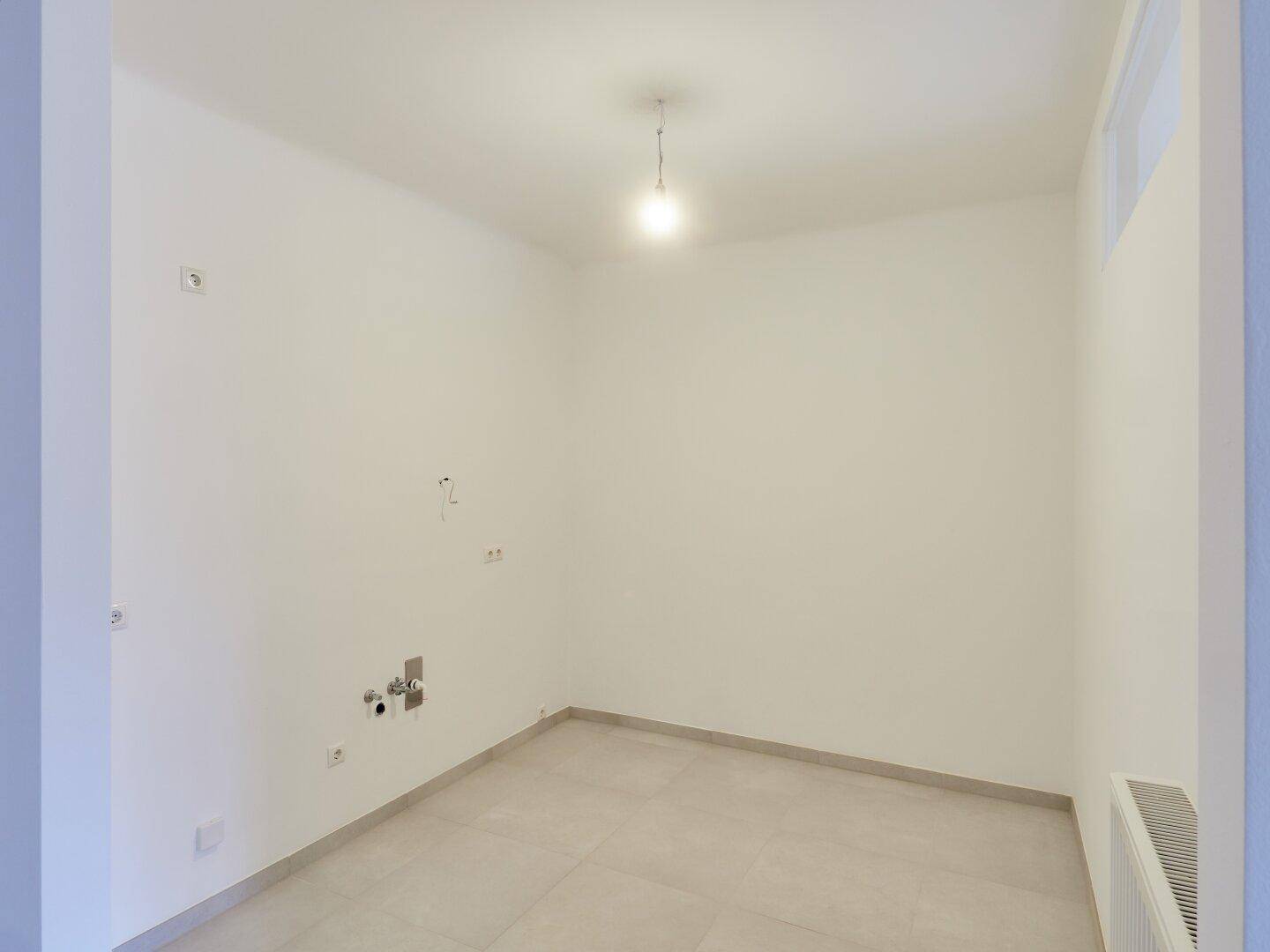 Heller Raum mit Fliesenboden und Wasseranschlüssen, bereit für die Installation einer Küche oder Waschküche.