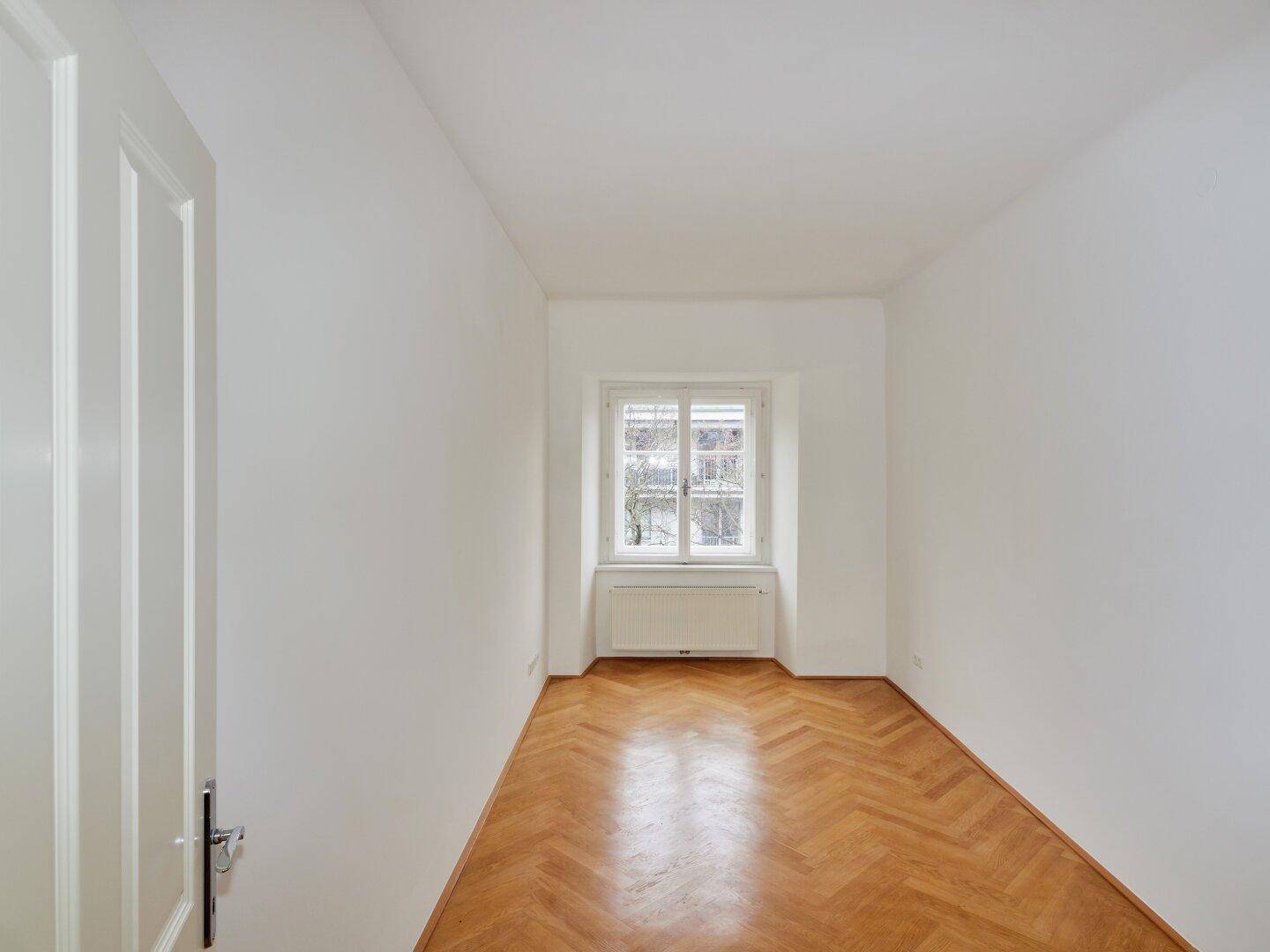 Langes, helles Zimmer mit Fischgrätparkett und einem Fenster, das den Raum mit Licht erfüllt.