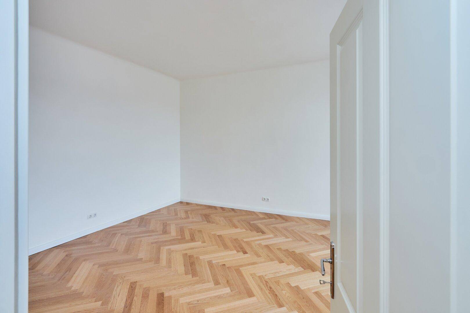 Leeres Zimmer mit hellem Fischgrätparkett und weiß gestrichenen Wänden, bereit zur Nutzung.