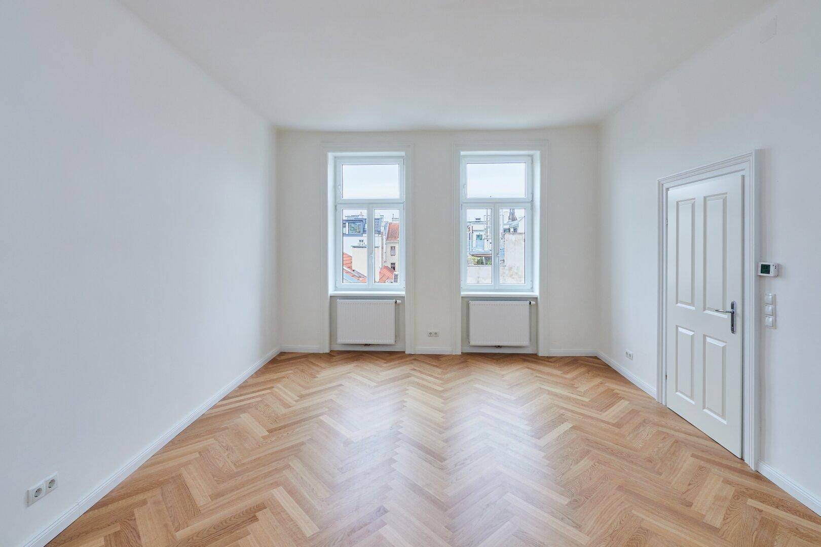 Geräumiges Zimmer mit klassischem Fischgrätparkett und weiß gestrichenen Wänden.