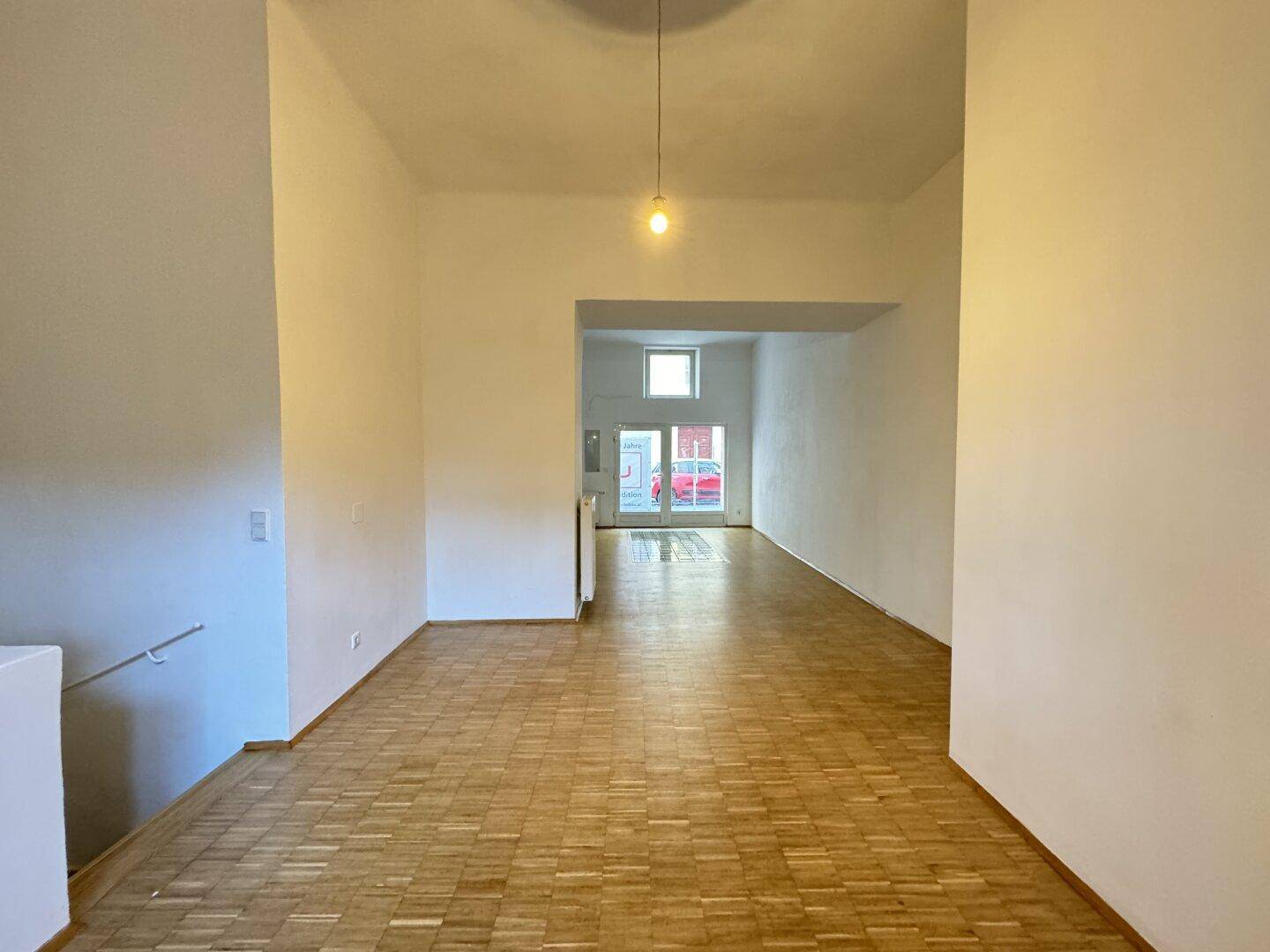 Langer, heller Raum mit Parkettboden und Blick auf die Straße durch ein Fenster im Hintergrund.