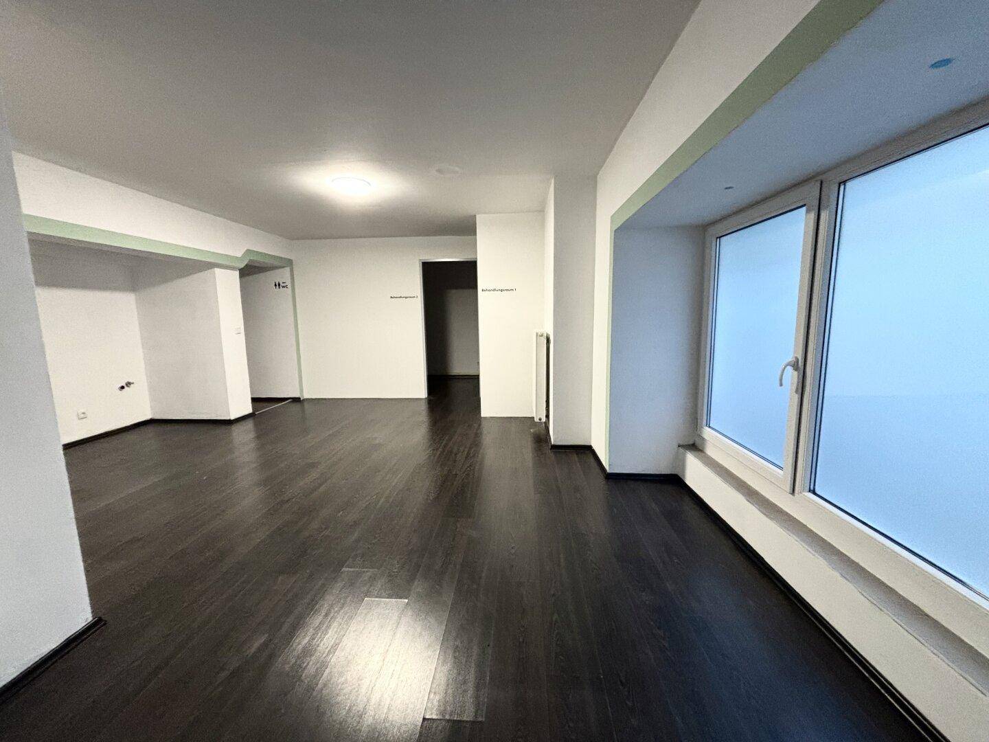 Großer, heller Raum mit dunklem Holzboden und breiten Fenstern, die viel Licht hereinlassen.
