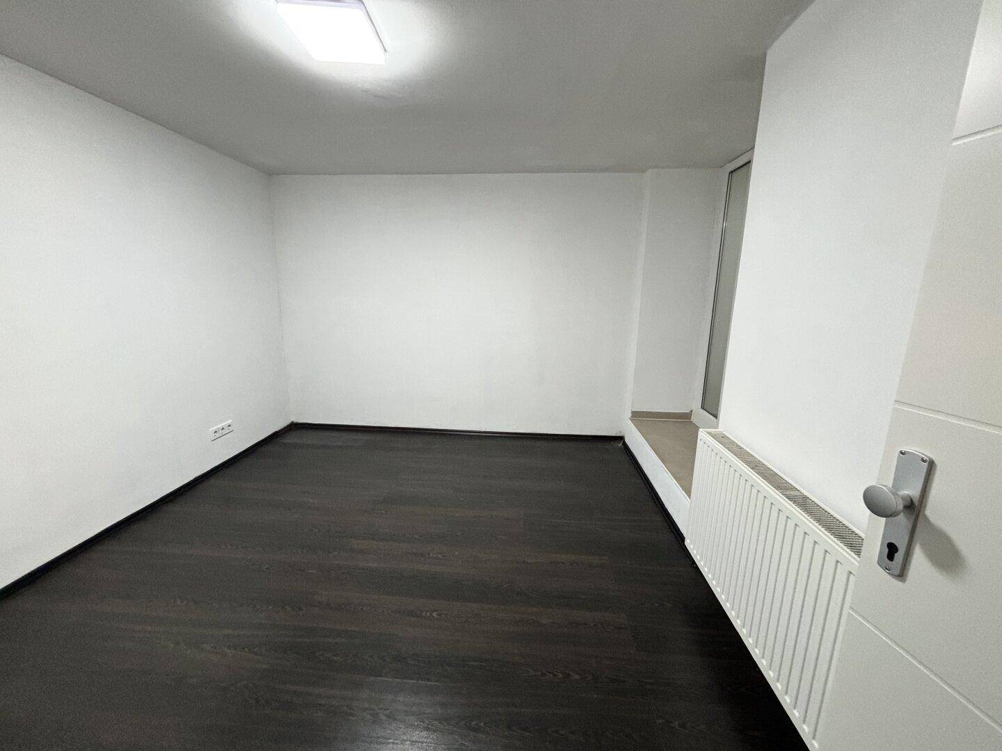 Kleiner, heller Raum mit dunklem Holzboden, einem Fenster und einem Heizkörper unterhalb.
