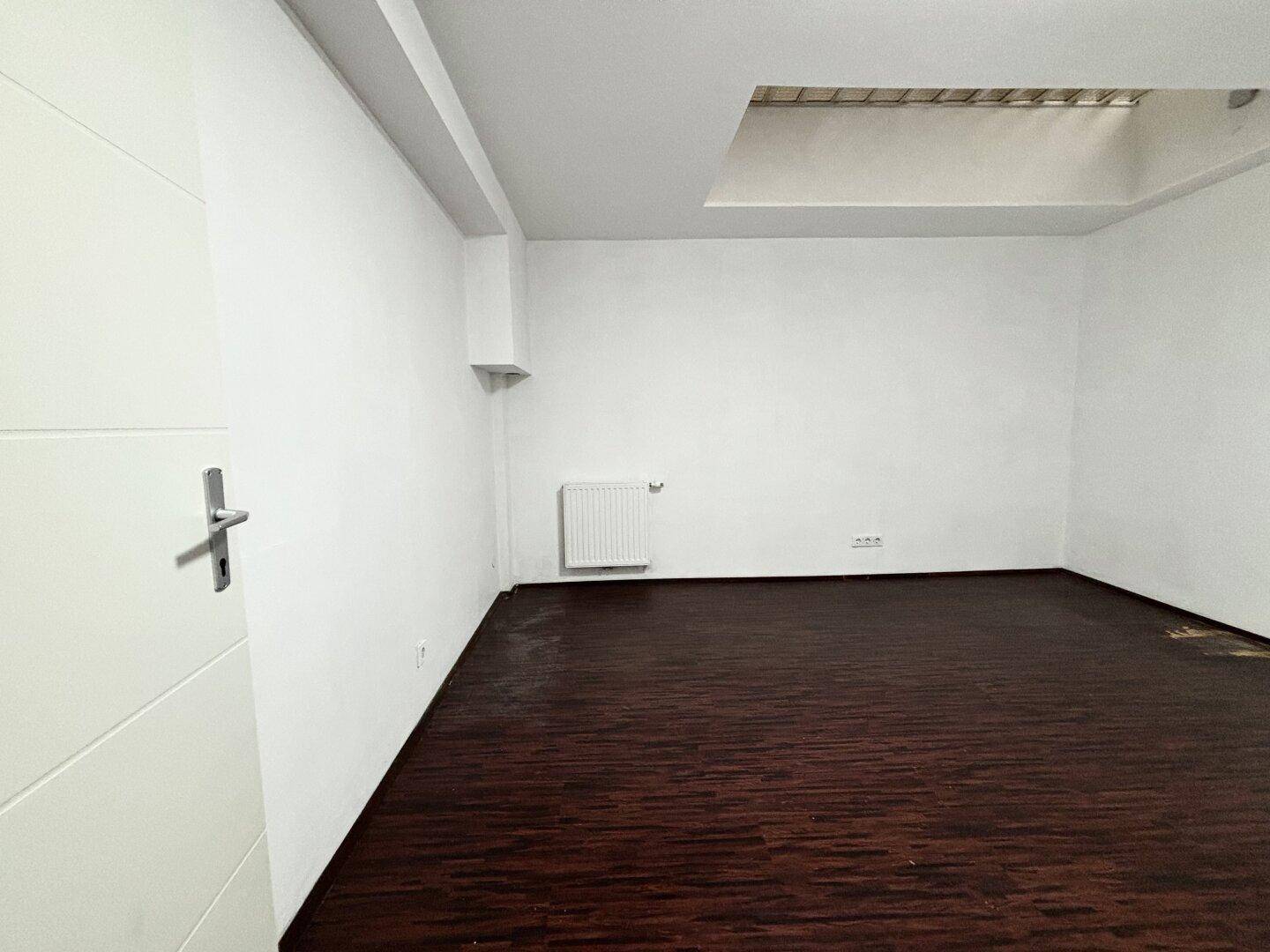 Heller Raum mit dunklem Holzboden, einem Heizkörper und einer Deckenöffnung mit Licht.