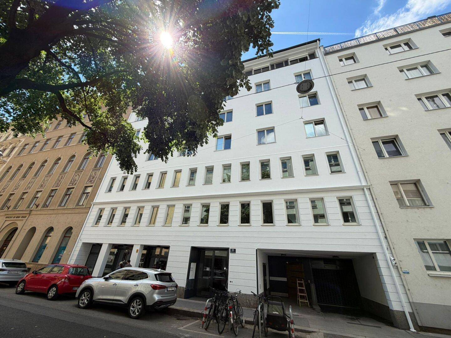 Moderne weiße Gebäudefassade an einer belebten Straße mit Bäumen und parkenden Autos.