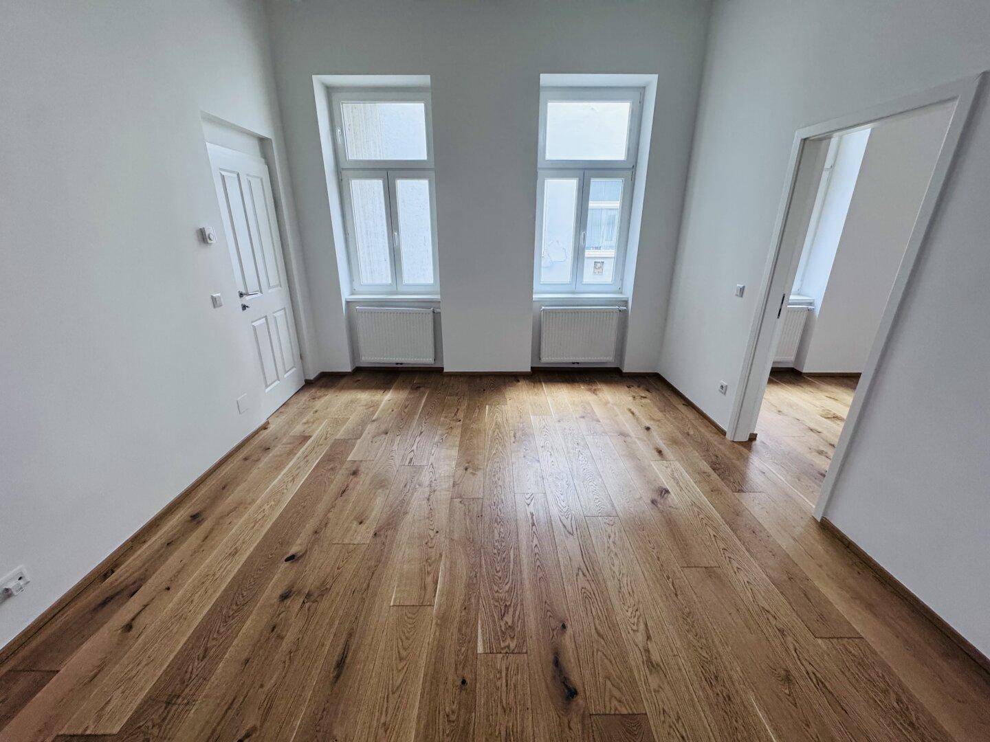 Leerer Raum mit Holzboden und zwei hohen Fenstern, bereit zur individuellen Gestaltung und Nutzung.