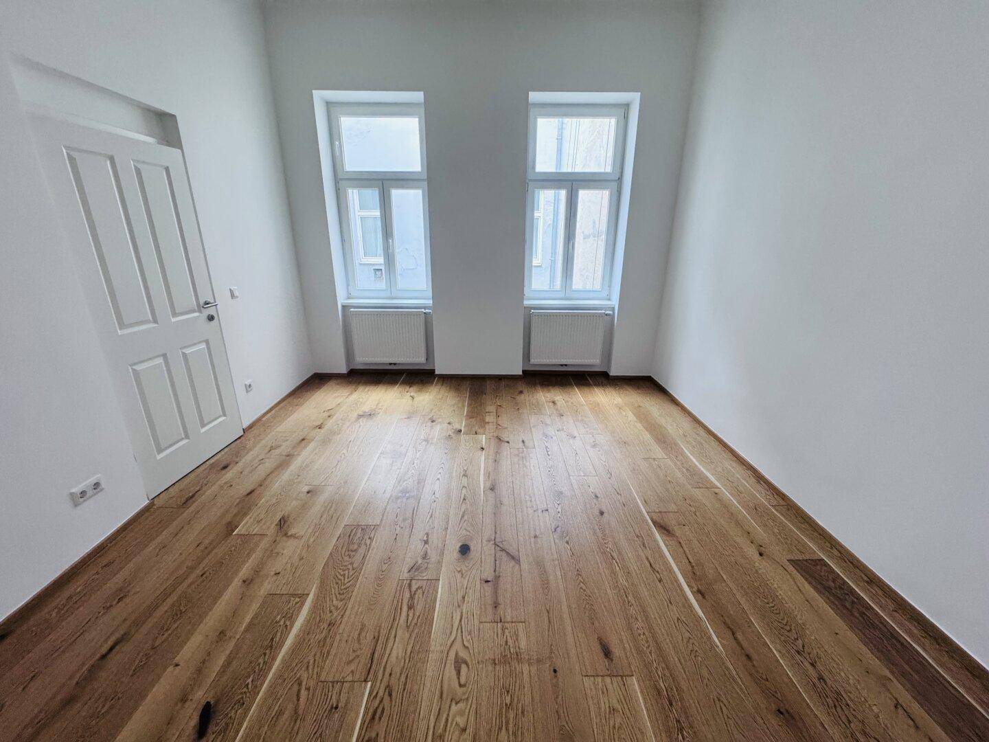 Heller, leerer Raum mit Holzboden und hohen Fenstern, ideal für Schlaf- oder Wohnbereich.