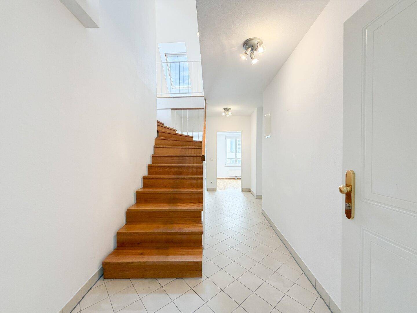 Heller Flur mit Holztreppe, Fliesenboden und einem Oberlicht für natürliches Licht.