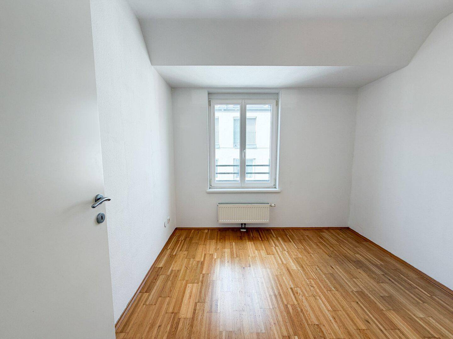 Ruhiges Zimmer mit Parkettboden, weißer Wandfarbe und einem großen Fenster.