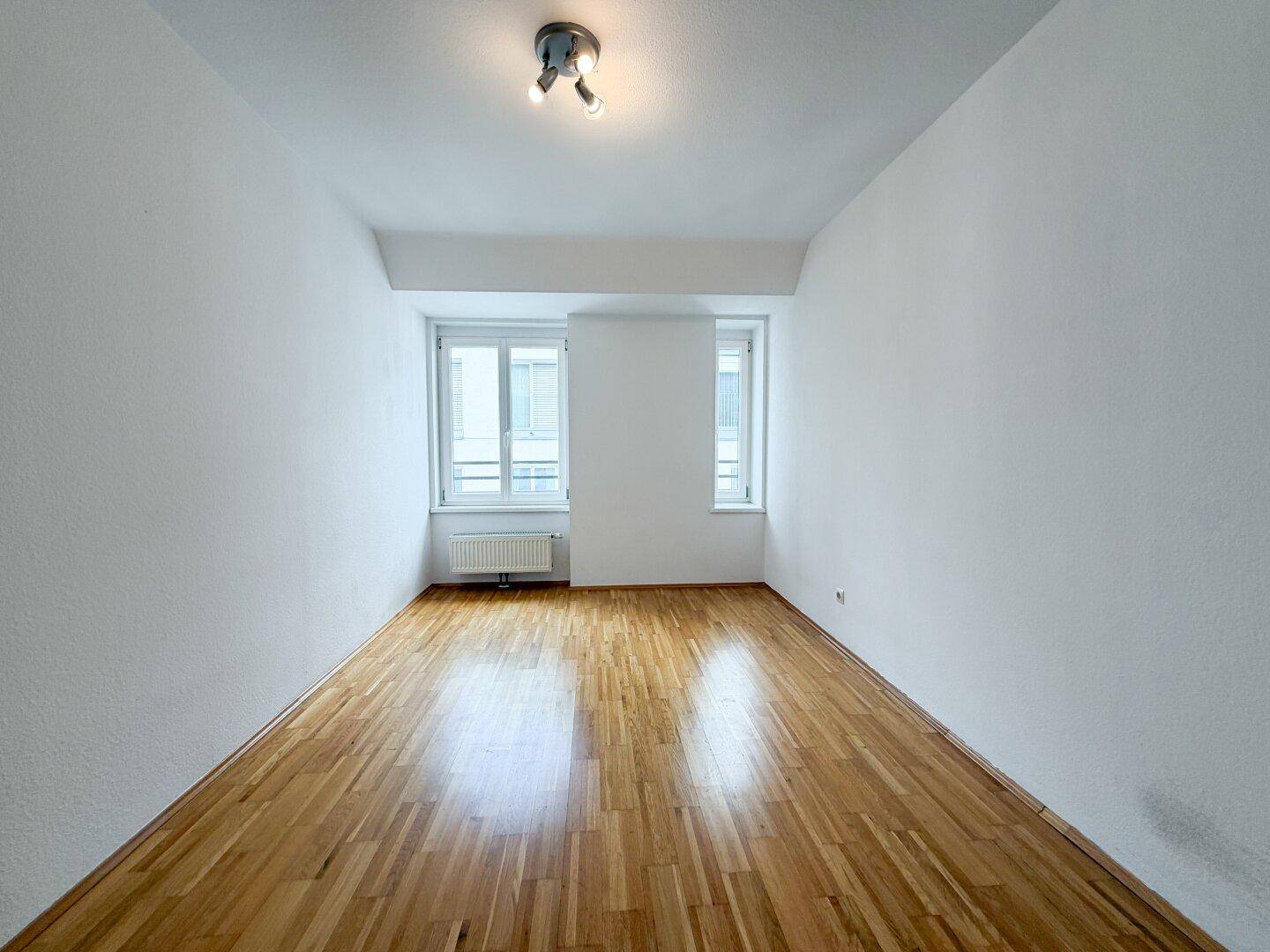 Leeres Zimmer mit hellem Parkettboden, weißen Wänden und zwei Fenstern.