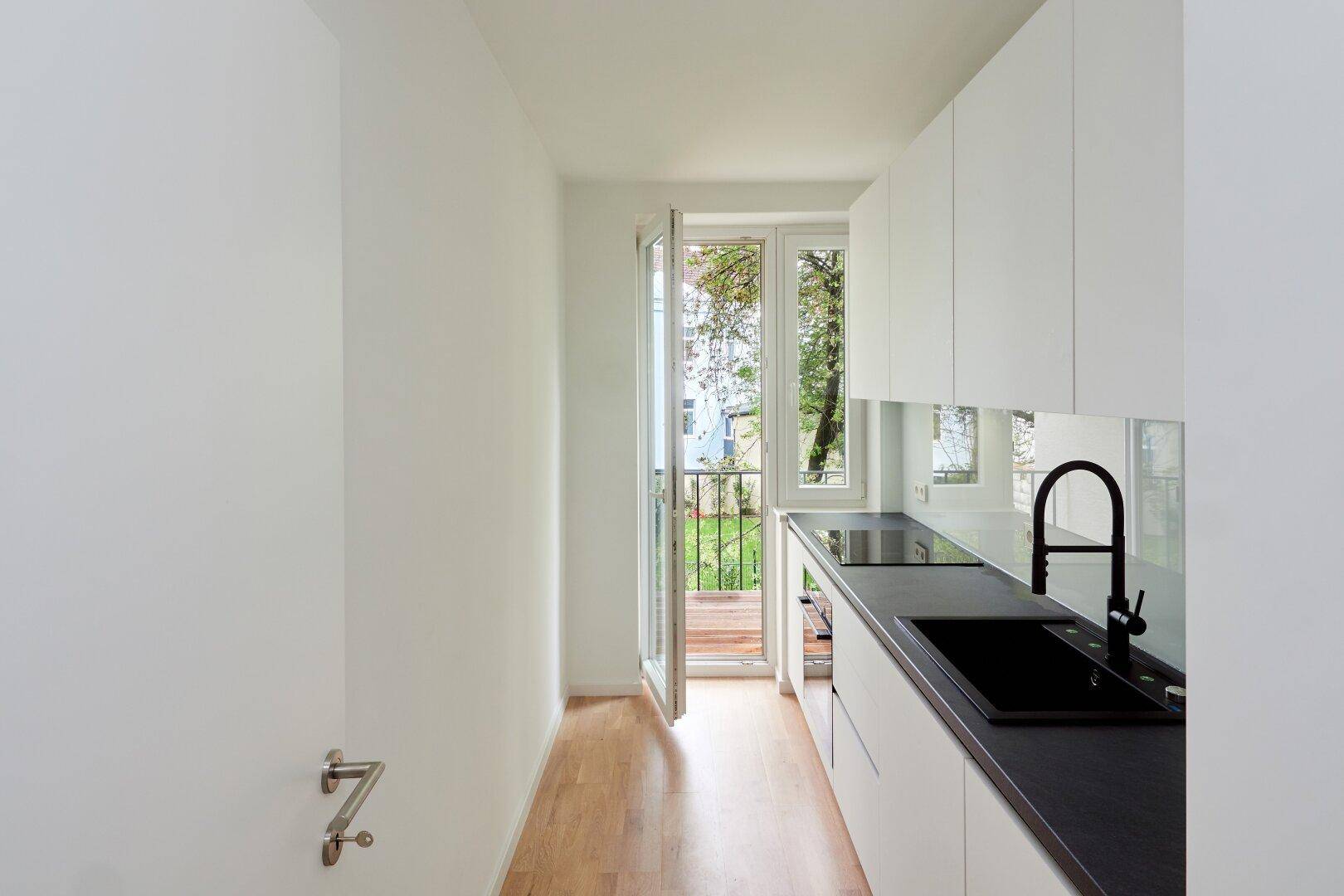Moderne Küche mit weißen Schränken, schwarzer Arbeitsplatte und Zugang zu einem Balkon mit Grünblick.