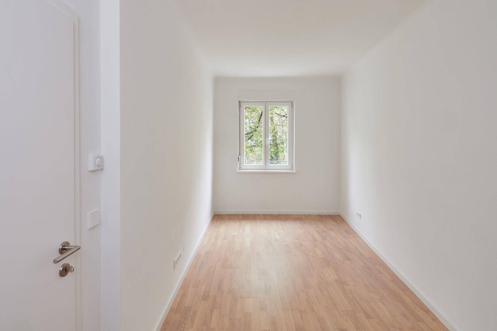 Gemütliches Zimmer mit Parkettboden und Fenster mit Blick auf Bäume und Grünflächen.