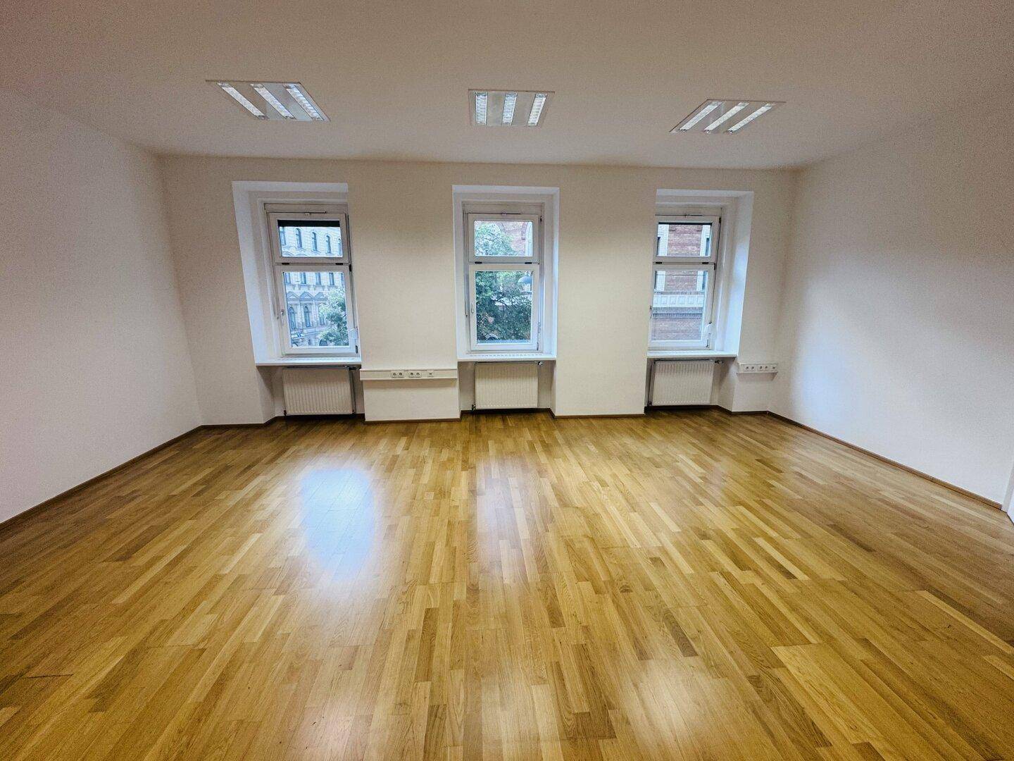 Heller Büroraum mit drei großen Fenstern und Holzparkettboden, bietet viel Tageslicht und eine angenehme Atmosphäre.