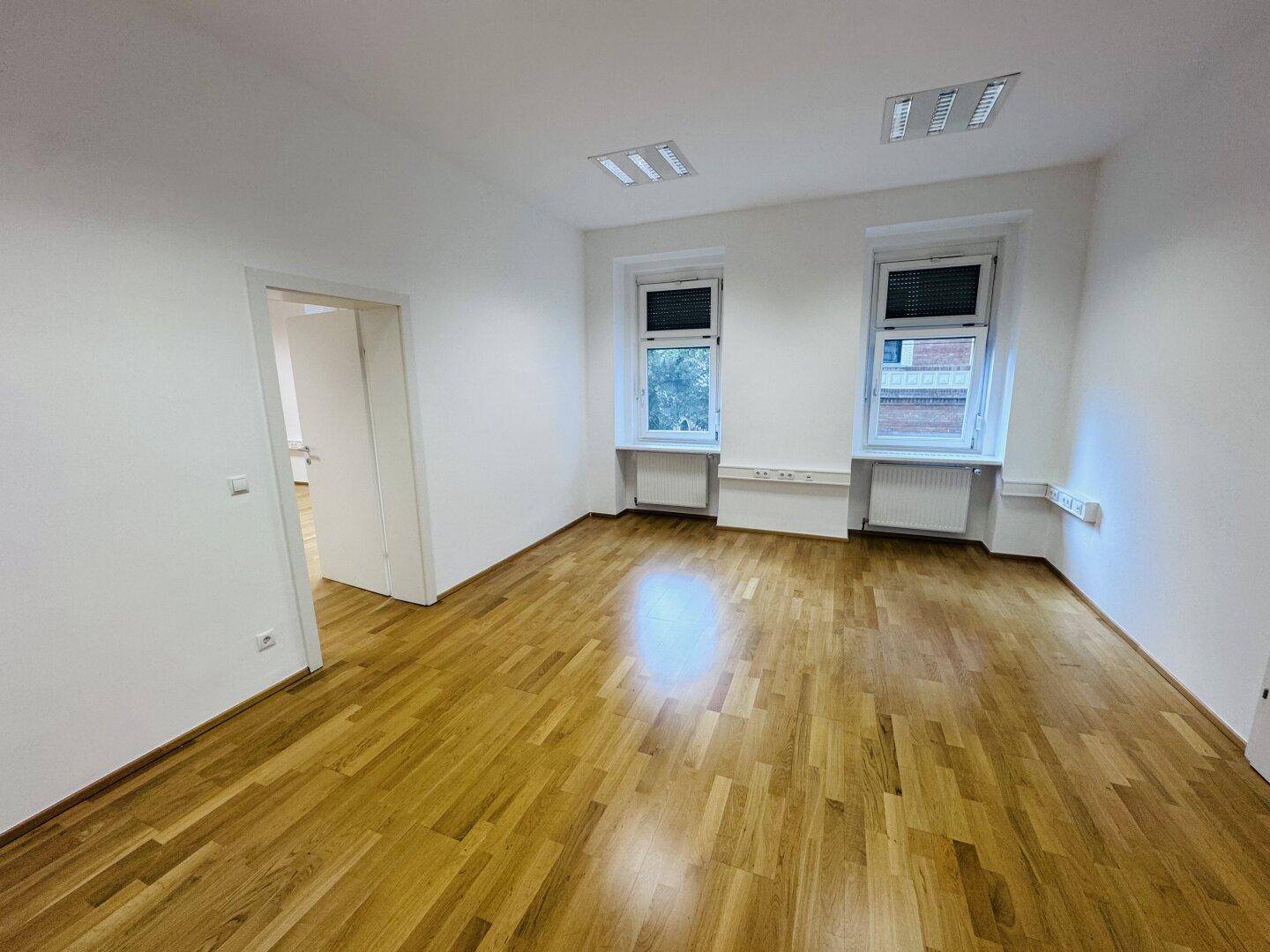 Leerer Büroraum mit zwei Fenstern und glänzendem Holzparkett, bereit für die individuelle Gestaltung.